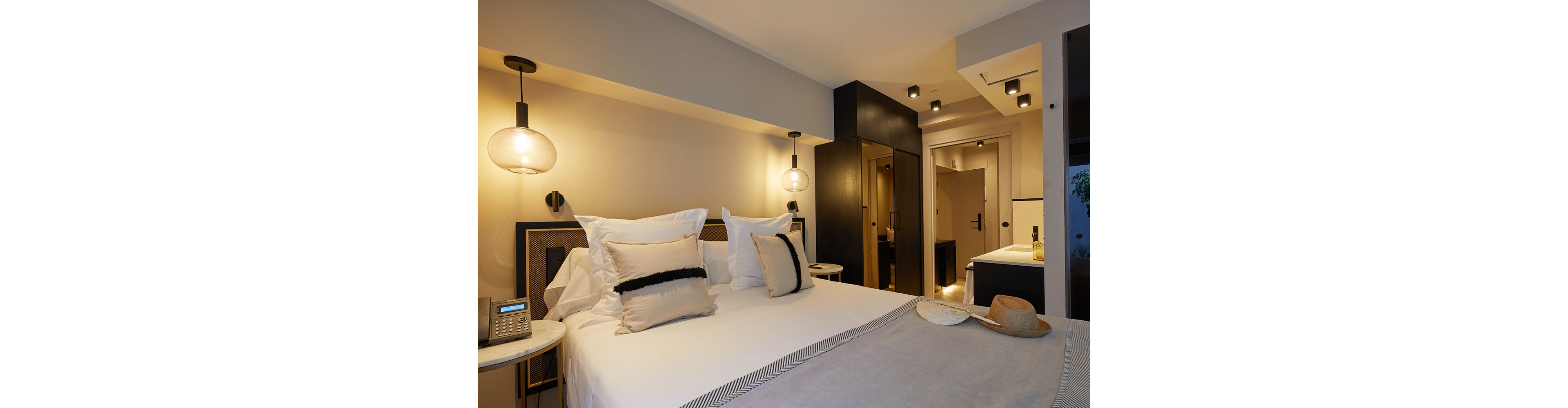 Contemporary hotel room interior featuring a neatly made double bed with layered pillows and a light throw blanket. Warm pendant lights and ceiling spotlights create a cozy atmosphere against neutral beige and black tones. A bedside table holds a telephone and hat, while built-in wardrobe and bathroom areas are visible in the background. The overall design emphasizes modern comfort and minimalist style.