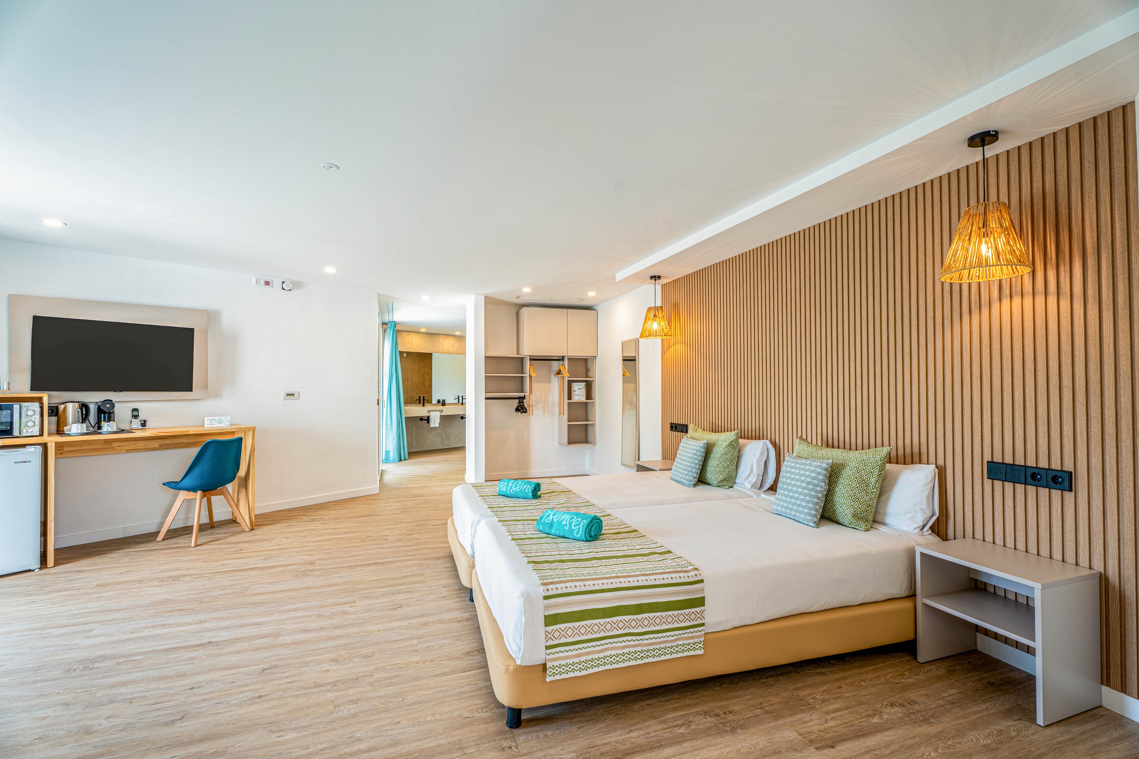 A spacious modern hotel room features a large double bed with striped green runners and neatly folded turquoise towels. The room includes a wooden desk area with a flat-screen television, mini fridge, and coffee station. Warm wooden flooring and a slatted accent wall complement woven pendant lights for a cozy yet contemporary feel. An open doorway reveals a bright bathroom or dressing area with additional storage and mirrors.