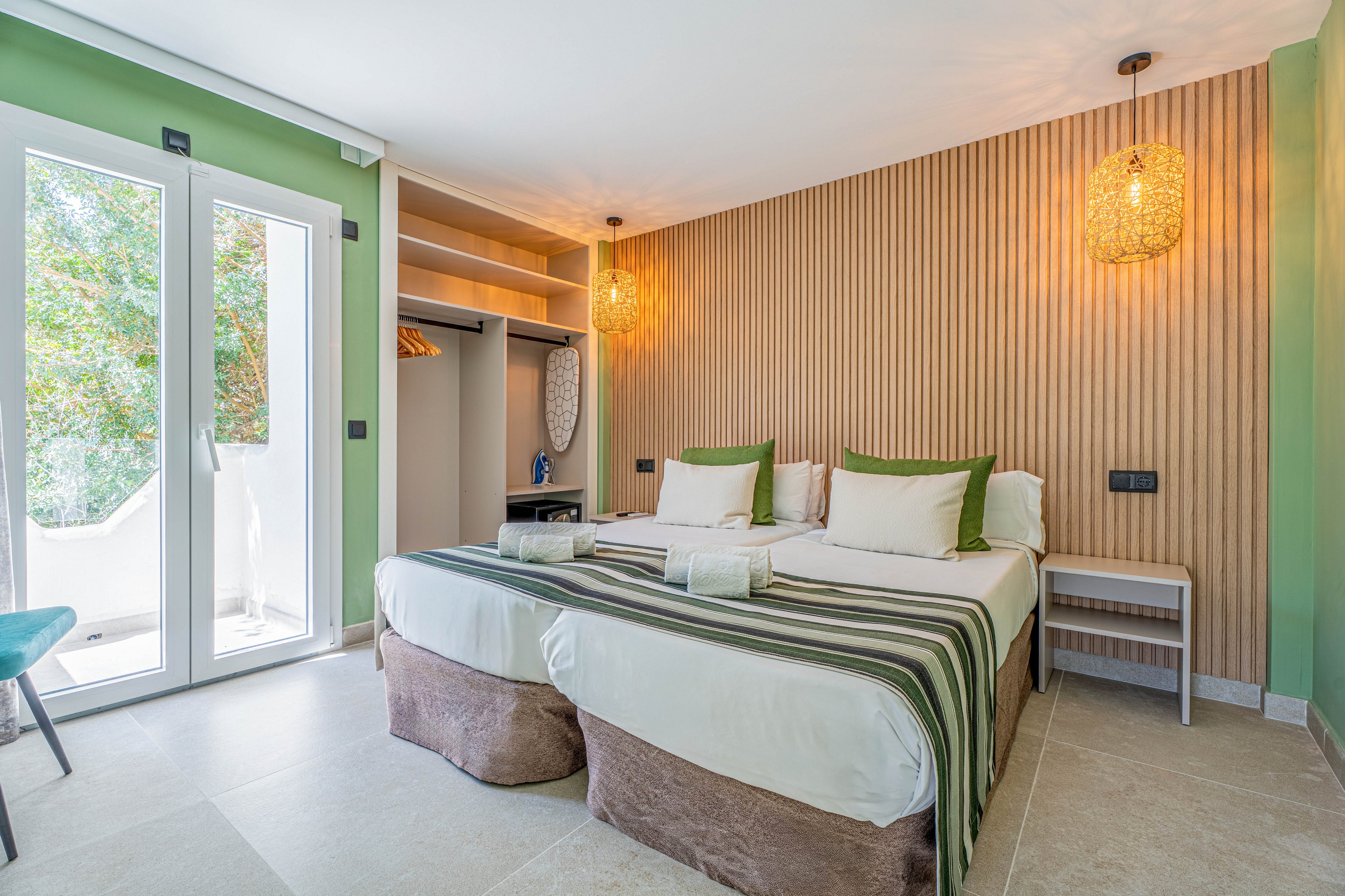 Bright contemporary bedroom featuring two neatly made twin beds with striped runners and green accent pillows. The room has a wooden slat feature wall, pendant lights, and soft neutral flooring. An open wardrobe with hangers and shelves is visible beside the beds. Large glass doors let in natural light and offer a view of greenery outside, creating an inviting and relaxing atmosphere.