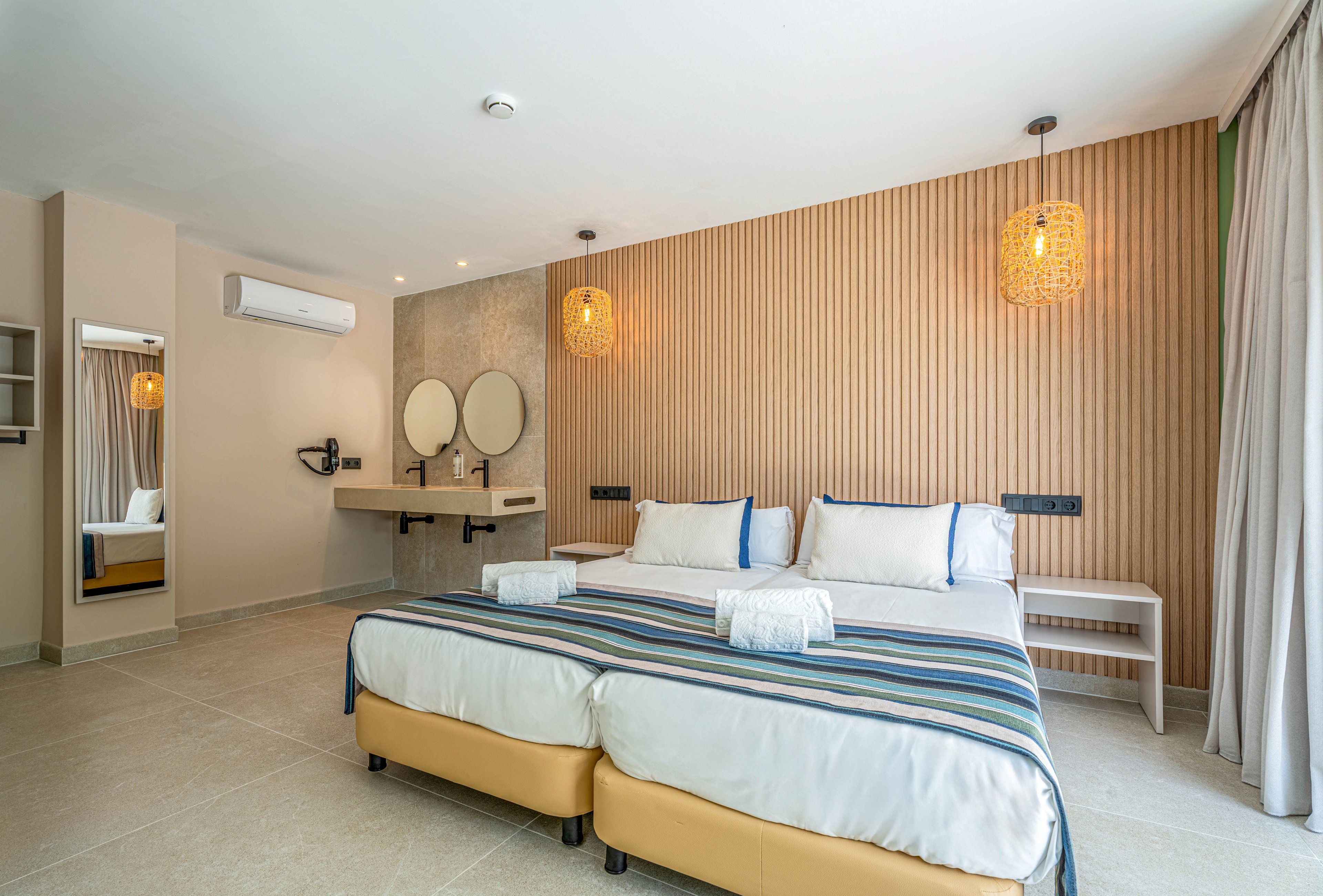 Spacious modern hotel room featuring two neatly made twin beds with striped bed runners and folded towels. The room includes a minimalist vanity area with round mirrors and wall-mounted lighting. Warm woven pendant lamps and a wood-paneled accent wall add texture and a cozy atmosphere. Large curtains cover a window or balcony door, allowing natural light to brighten the neutral-toned interior.