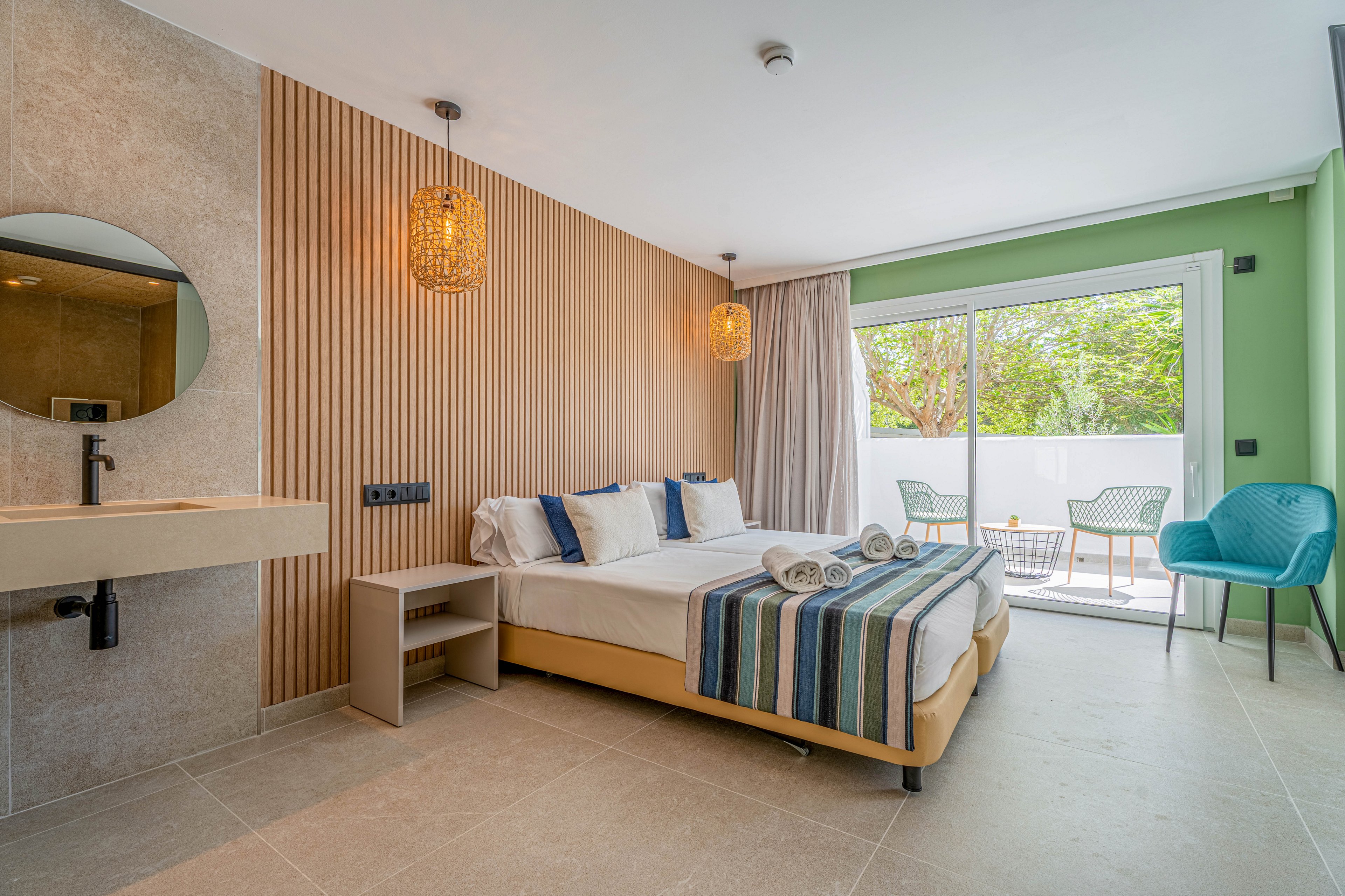 Contemporary hotel room featuring a double bed neatly made with striped throws and rolled towels. The space includes a built-in sink area, bedside shelving, and warm wooden slat accent wall with hanging woven lights. Large sliding glass doors open to a bright terrace with outdoor chairs and greenery visible outside. Neutral floor tiles and a teal accent chair add to the clean, modern aesthetic.