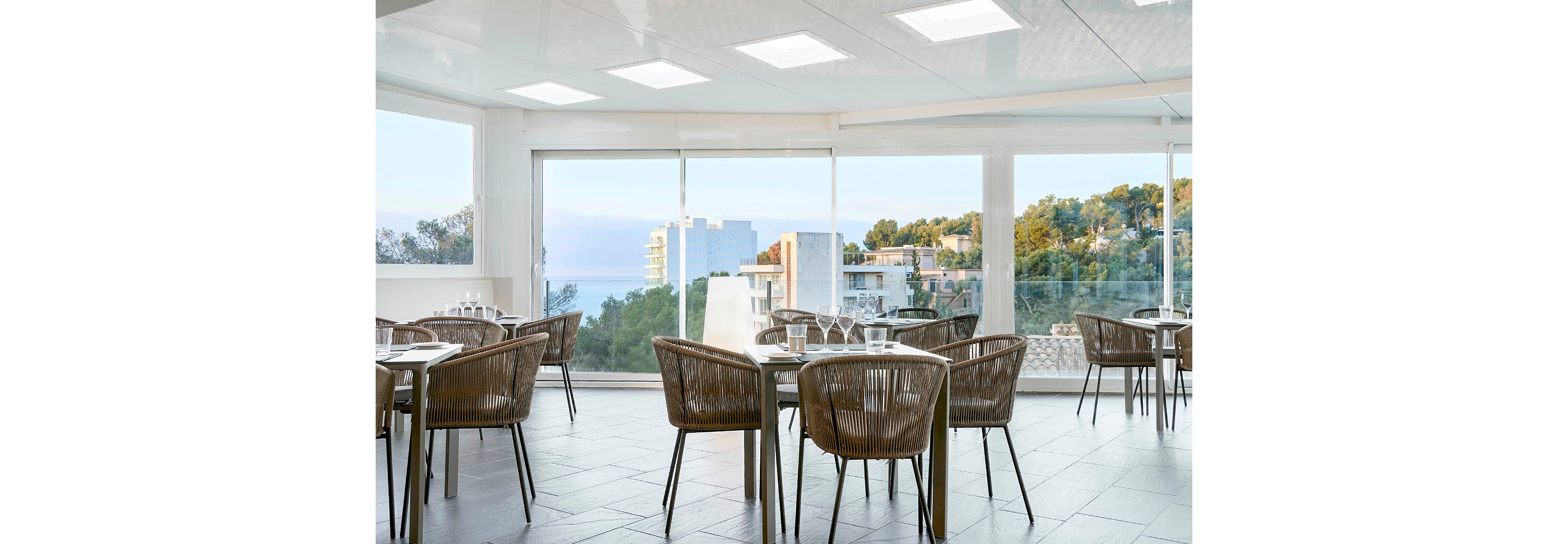 A bright, modern restaurant dining area features several empty tables and woven chairs neatly arranged. Large floor-to-ceiling windows reveal a coastal town with trees, buildings, and a distant view of the sea. Natural light fills the space, reflecting off the tiled floor and clean white ceiling panels. The scene feels calm and inviting, suggesting a relaxed dining experience with scenic views.
