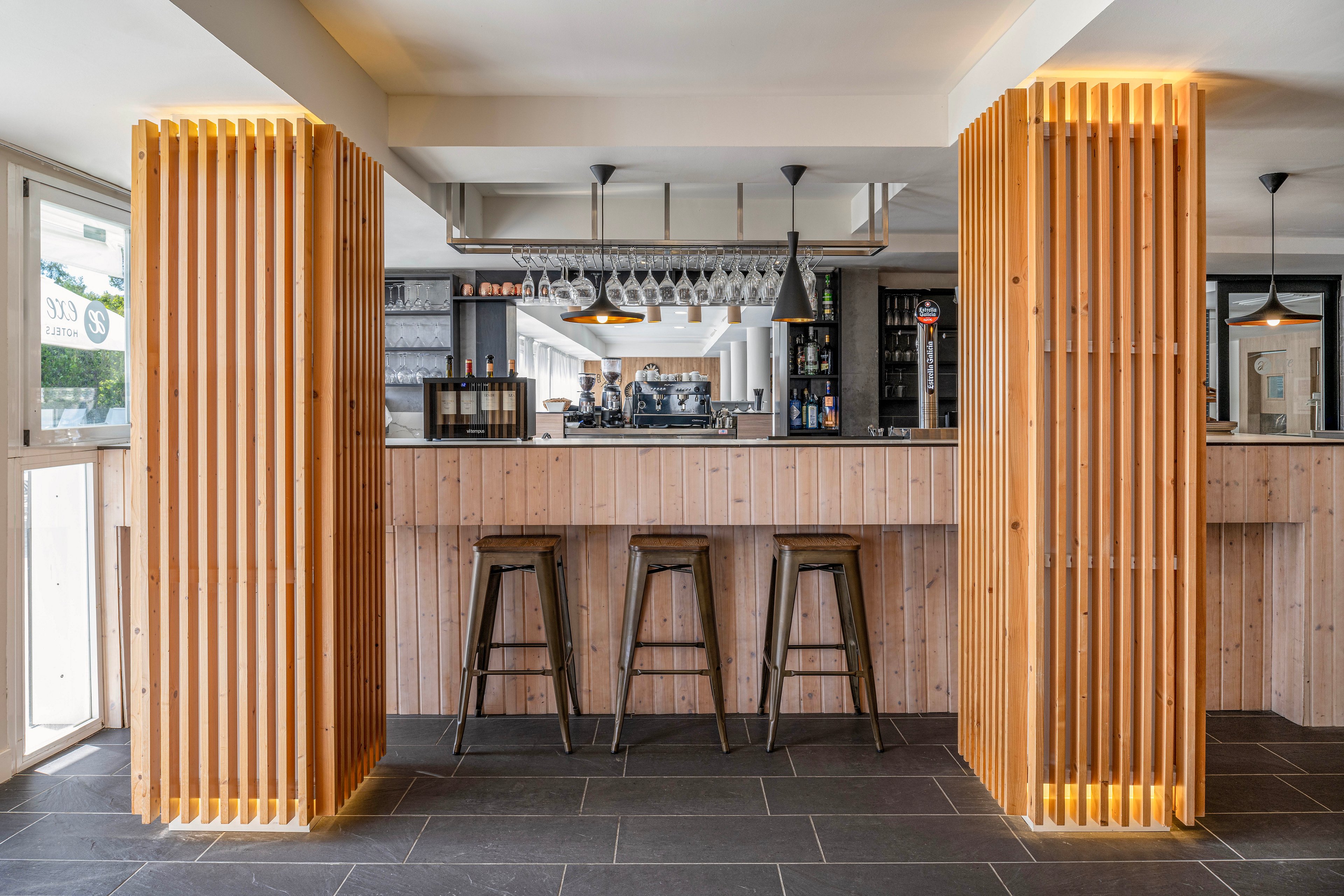 Contemporary bar interior featuring a light wood counter and vertical wooden slats with integrated lighting. Three metal bar stools are lined up in front of the bar, facing the shelves and coffee machine in the background. Hanging glassware and pendant lights add a stylish touch above the counter. The space is framed by large wooden columns and dark floor tiles, creating a clean, minimalist atmosphere.