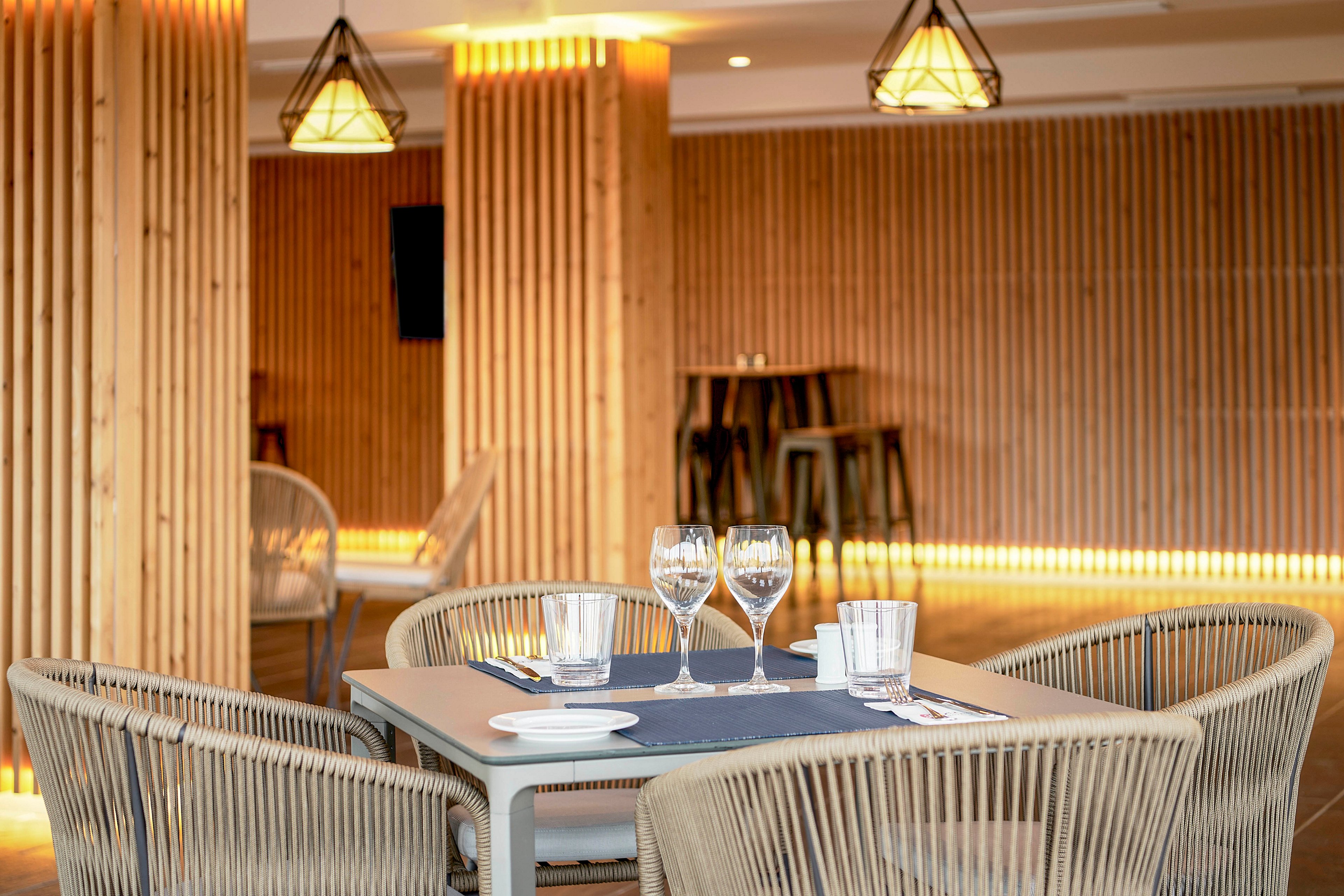 A neatly set dining table stands in a modern restaurant interior. The table features empty wine glasses, water glasses, and a small white plate on a blue runner. Woven chairs surround the table, adding a contemporary touch. In the background, wooden slat walls, warm lighting, and hanging pendant lamps create a cozy, minimalist atmosphere.