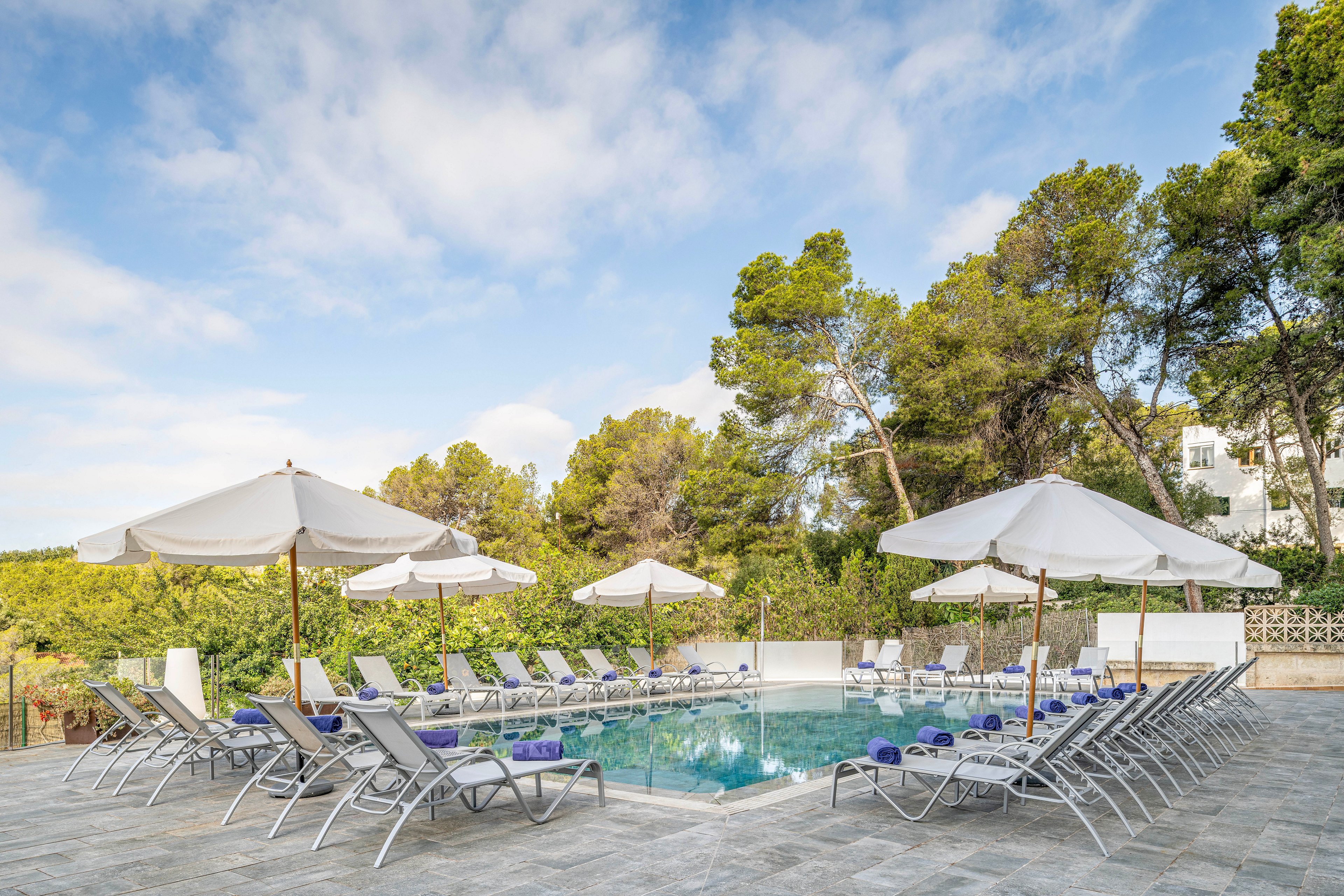 An outdoor swimming pool is surrounded by neatly arranged sun loungers and large white parasols. The pool area is set on a tiled terrace bordered by green trees and shrubs. Rolled blue towels rest on many of the loungers, suggesting a prepared but currently empty resort space. The scene is lit by soft daylight under a partly cloudy blue sky, creating a calm vacation atmosphere.