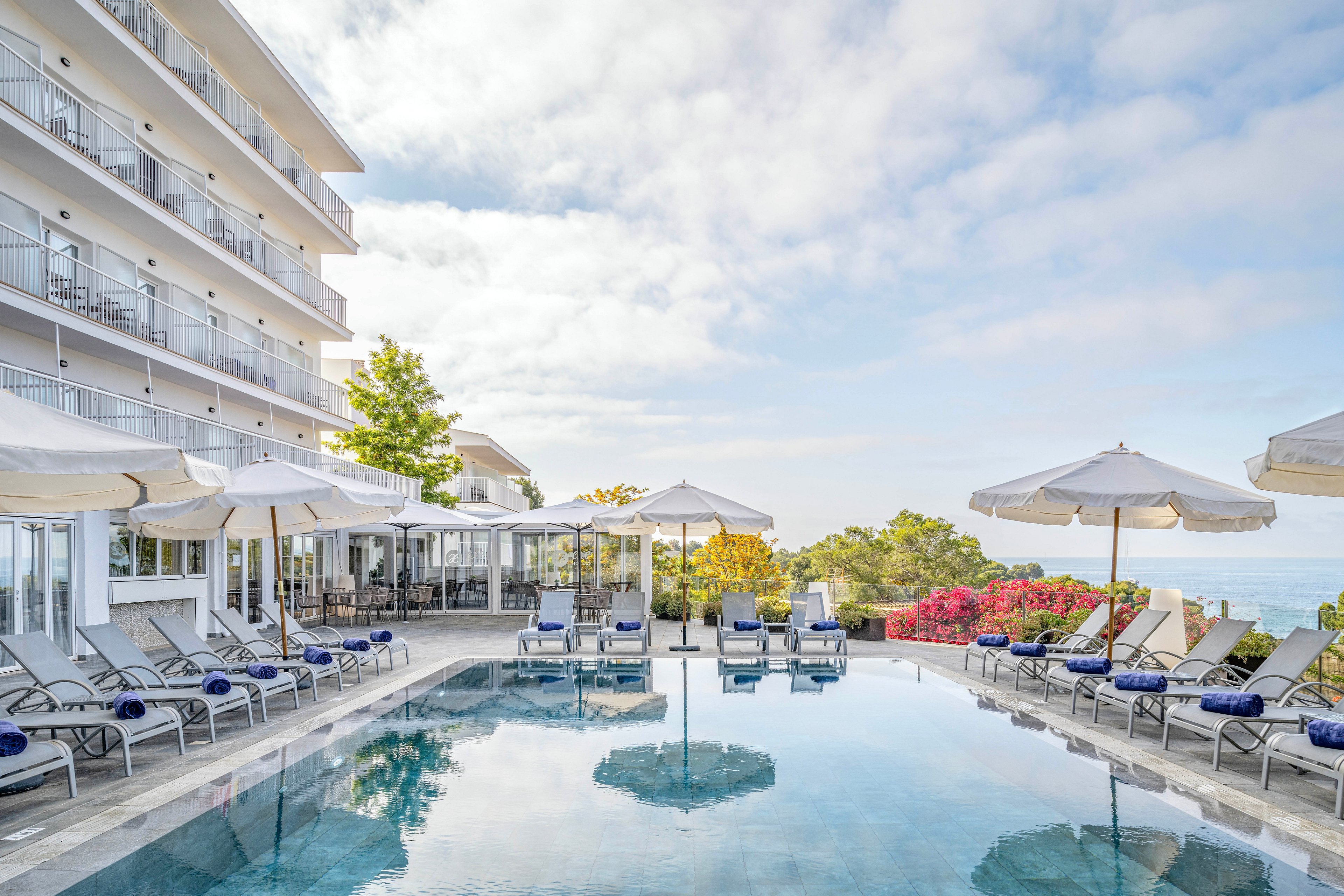 An upscale hotel features a serene outdoor swimming pool lined with lounge chairs and umbrellas. The poolside terrace overlooks a distant ocean horizon under a partly cloudy sky. White buildings with balconies frame one side, while colorful flowering shrubs add contrast near the edge. The calm water reflects the surrounding architecture and umbrellas, creating a tranquil vacation atmosphere.