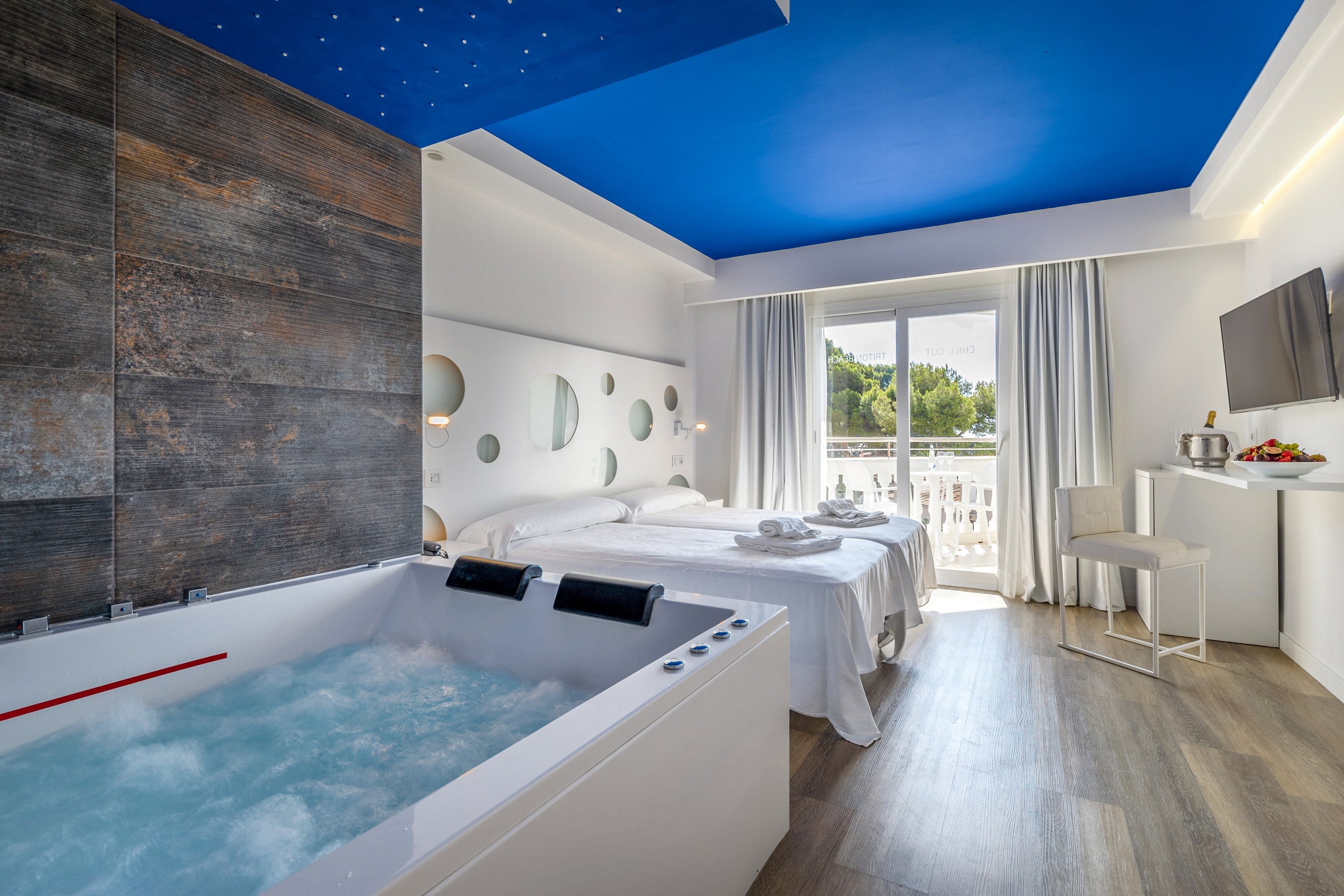 A contemporary hotel room featuring a jacuzzi with bubbling water in the foreground. The setting includes a double bed, a balcony with outdoor views, and minimalist decor. The ceiling is painted bright blue, complementing the neutral tones of the room. The space is well-lit with natural light streaming through the balcony doors.