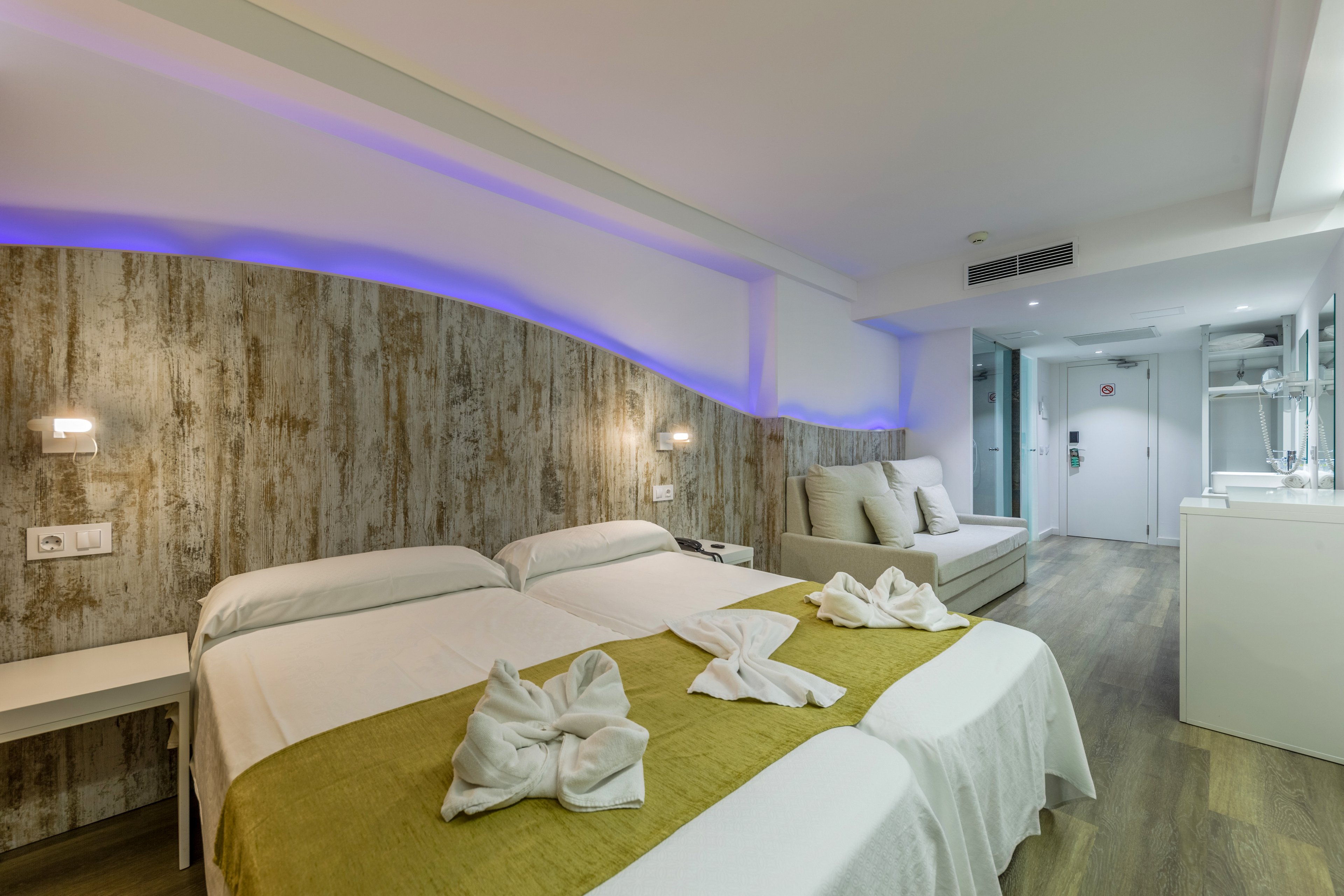 A contemporary hotel room featuring twin beds with neatly folded towel decorations on a mustard-colored runner. The room has a rustic wooden accent wall with soft purple lighting, a seating area, and a clean, minimalist design. The flooring is wooden, and the space is well-lit with modern fixtures.