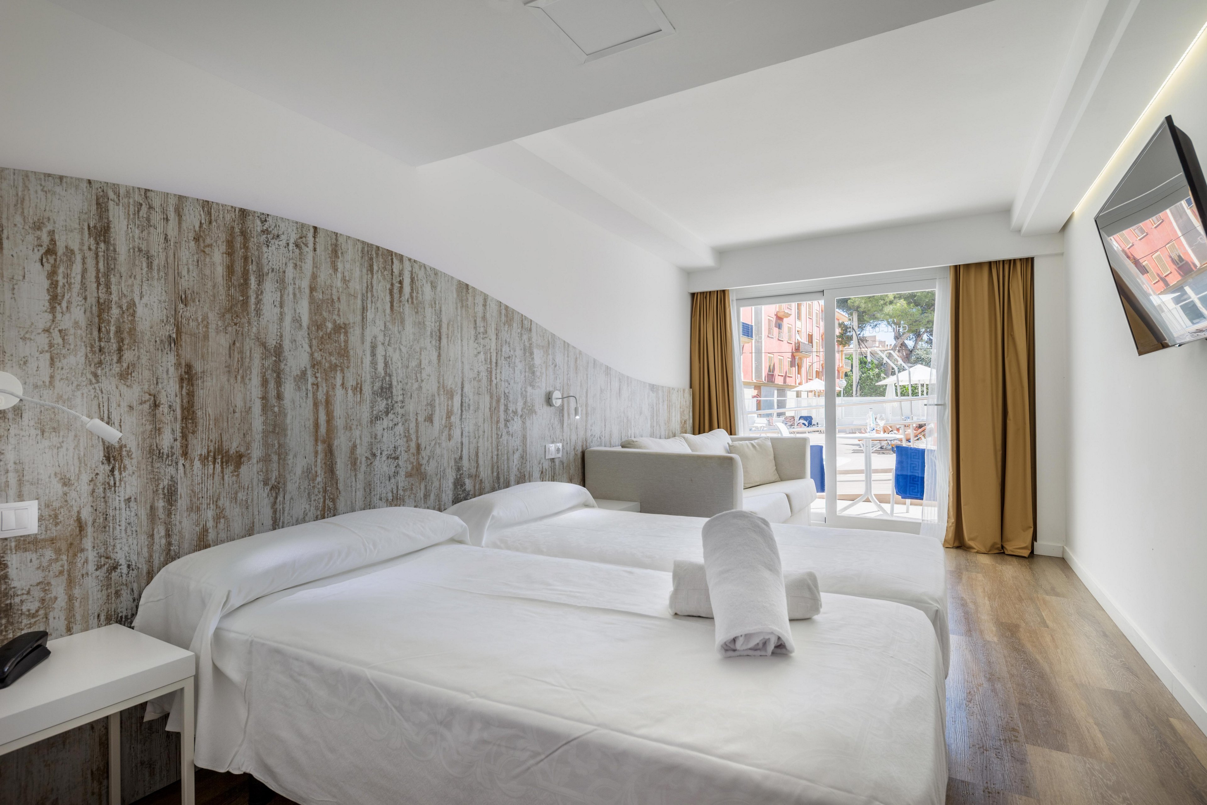 A spacious hotel room featuring two neatly made beds and a sofa, with a rustic wooden accent wall. The room opens to a balcony with a view of colorful buildings and greenery. Bright natural light fills the space, complemented by warm tones and minimalist decor.