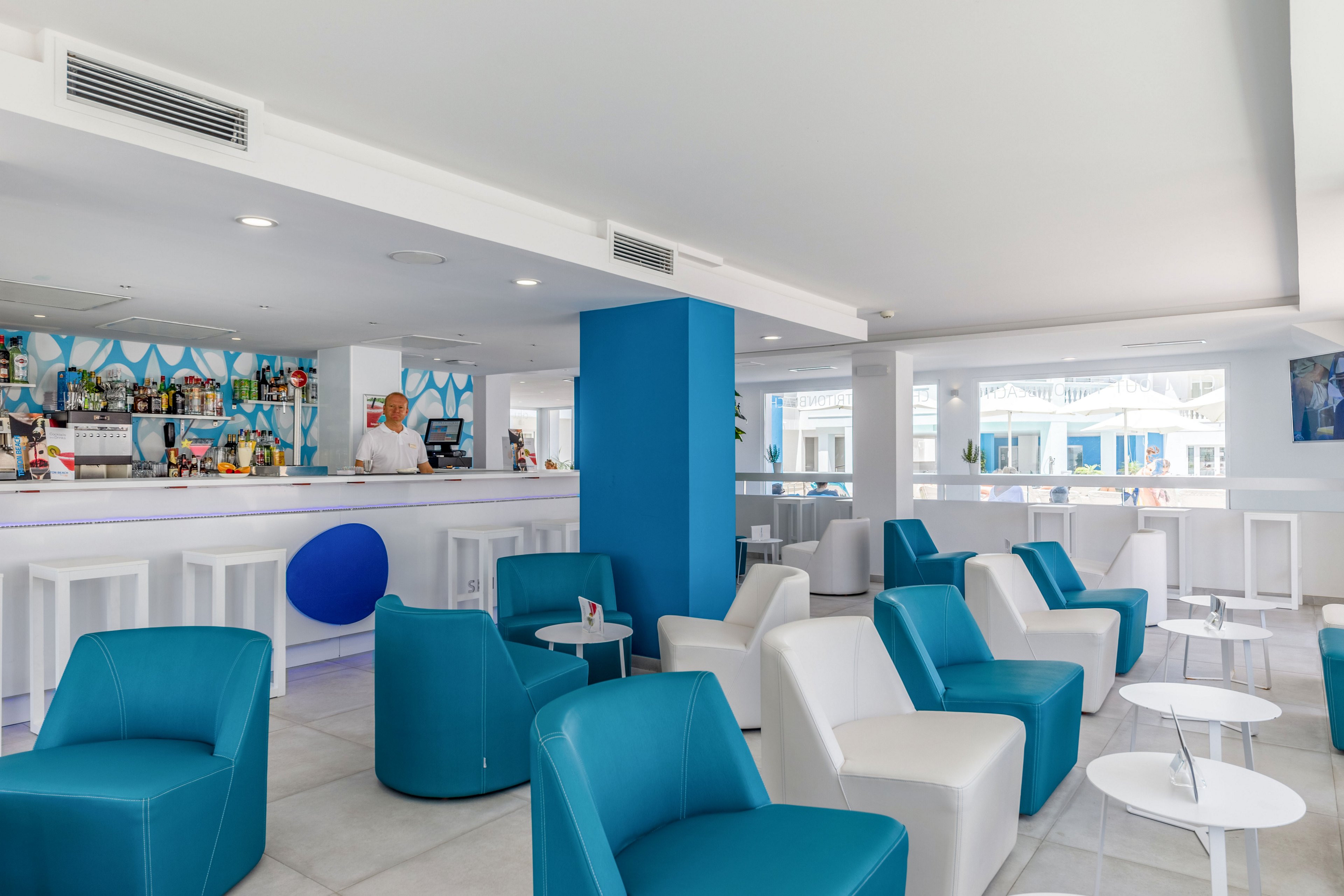 Contemporary lounge bar interior with white and blue armchairs and small round tables. A bartender stands behind the brightly lit bar, which features colorful bottles and decor. Large windows and white walls create an airy, minimalist atmosphere with clean lines. The overall setting suggests a stylish hotel or resort relaxation area.