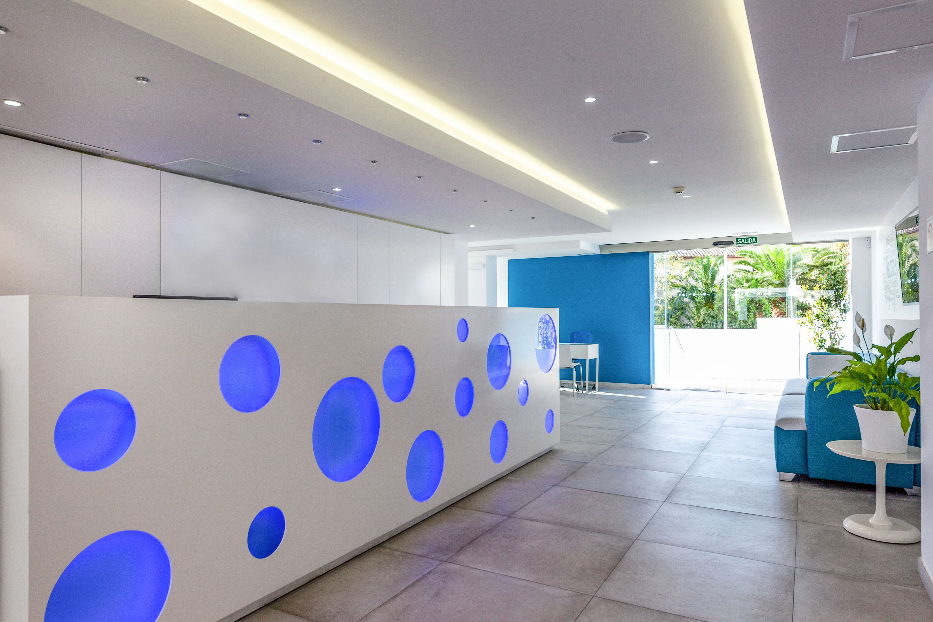 Contemporary reception desk with circular blue light insets stands in a bright, minimalist lobby. The space features clean white walls, large floor tiles, and a sleek ceiling light strip. A blue accent wall and seating area add color near the glass entrance, which opens to greenery outside. A small side table with a plant and chair completes the welcoming, modern interior.