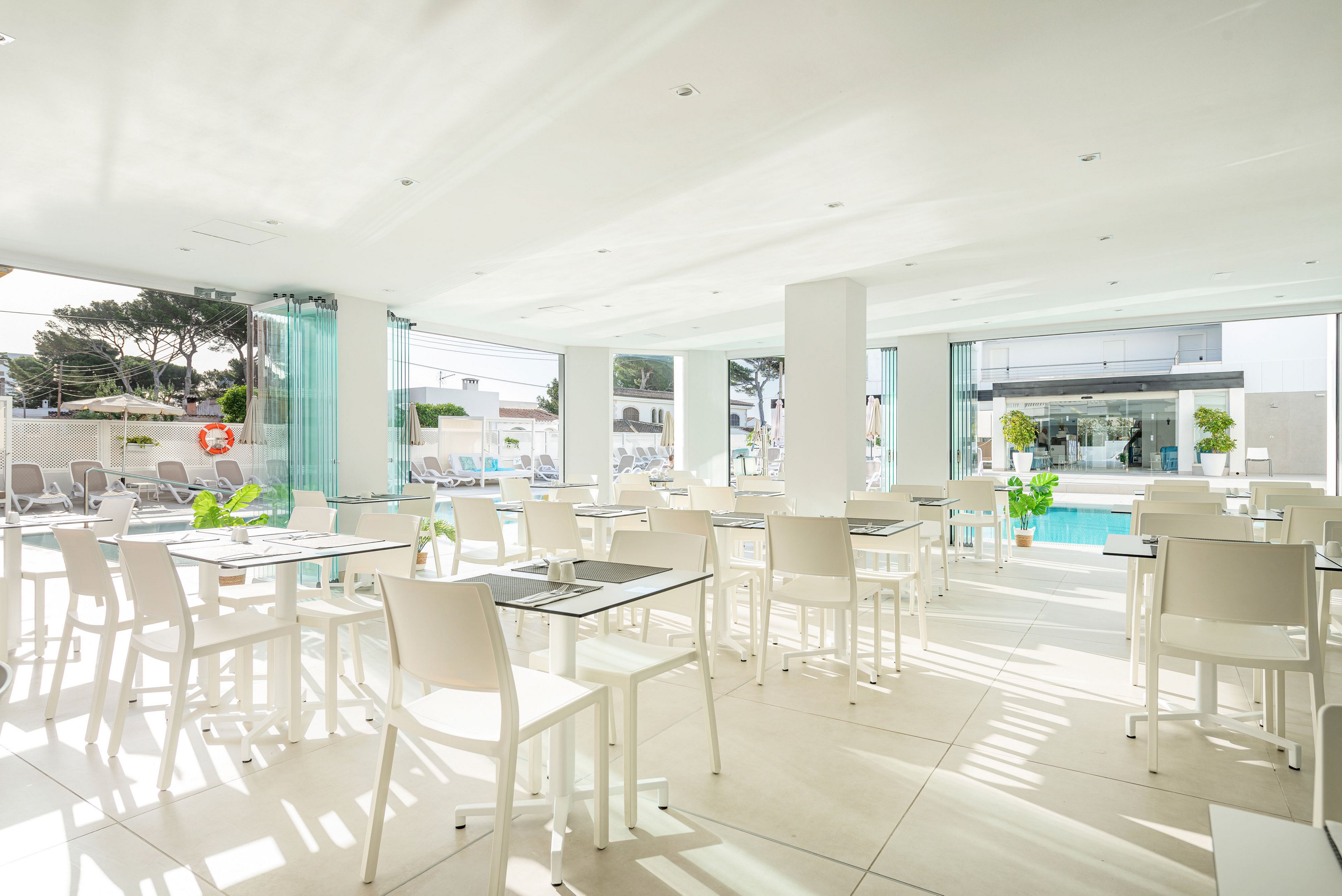 Spacious, contemporary dining area with white tables and chairs opens onto an outdoor swimming pool. Large floor-to-ceiling windows and glass panels let in abundant natural light, creating an airy atmosphere. Sun loungers and umbrellas are visible outside, suggesting a resort or hotel setting. Green plants and minimalist decor contribute to a clean, relaxed, vacation feel.