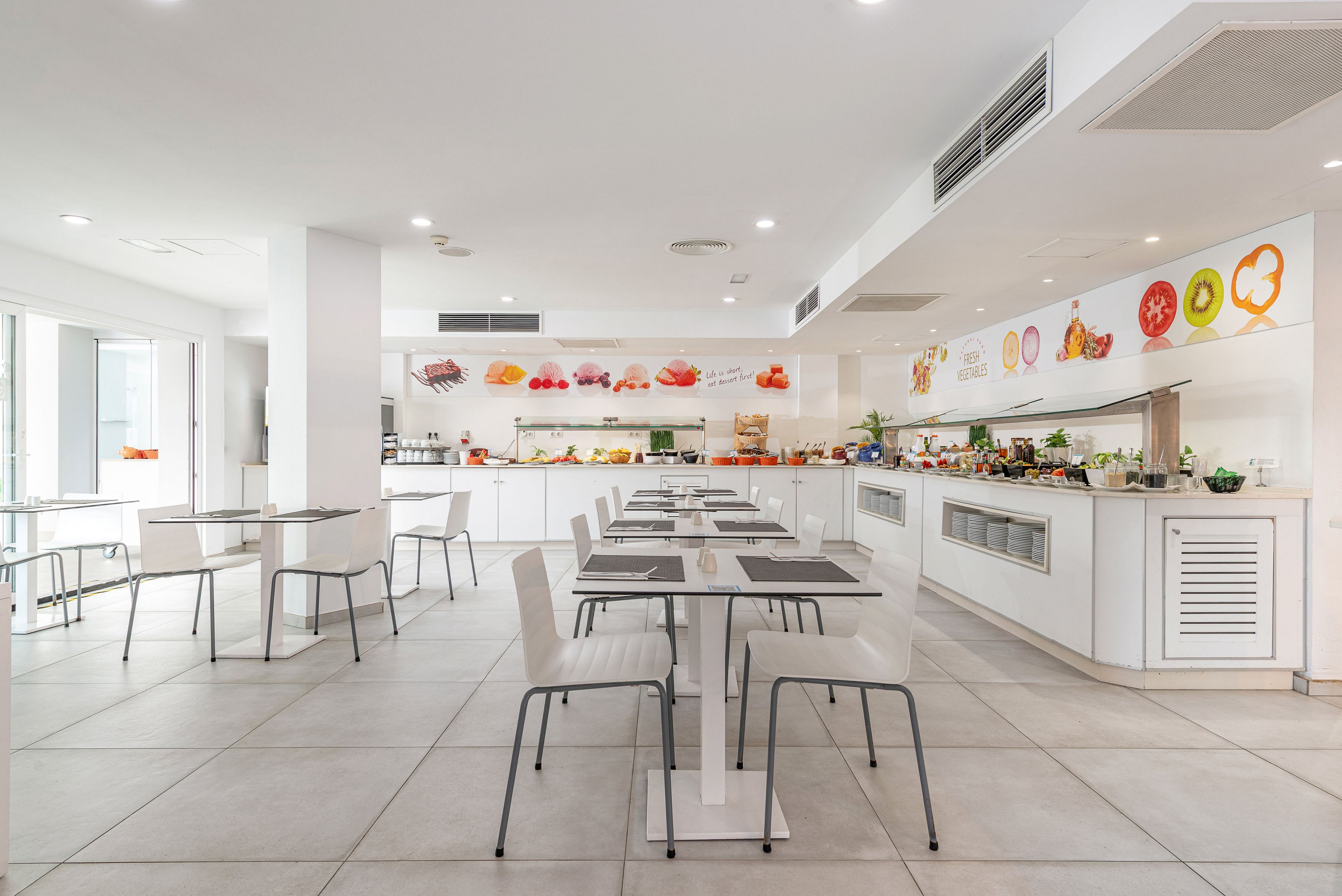 A spacious, modern cafeteria interior features rows of white tables and chairs arranged neatly on a light tiled floor. At the back, a long buffet counter displays various foods and beverages under bright lighting. Colorful fruit and food illustrations decorate the wall above the counter, adding visual interest to the clean white space. Large windows on the side bring in natural light, enhancing the airy and minimalist atmosphere.