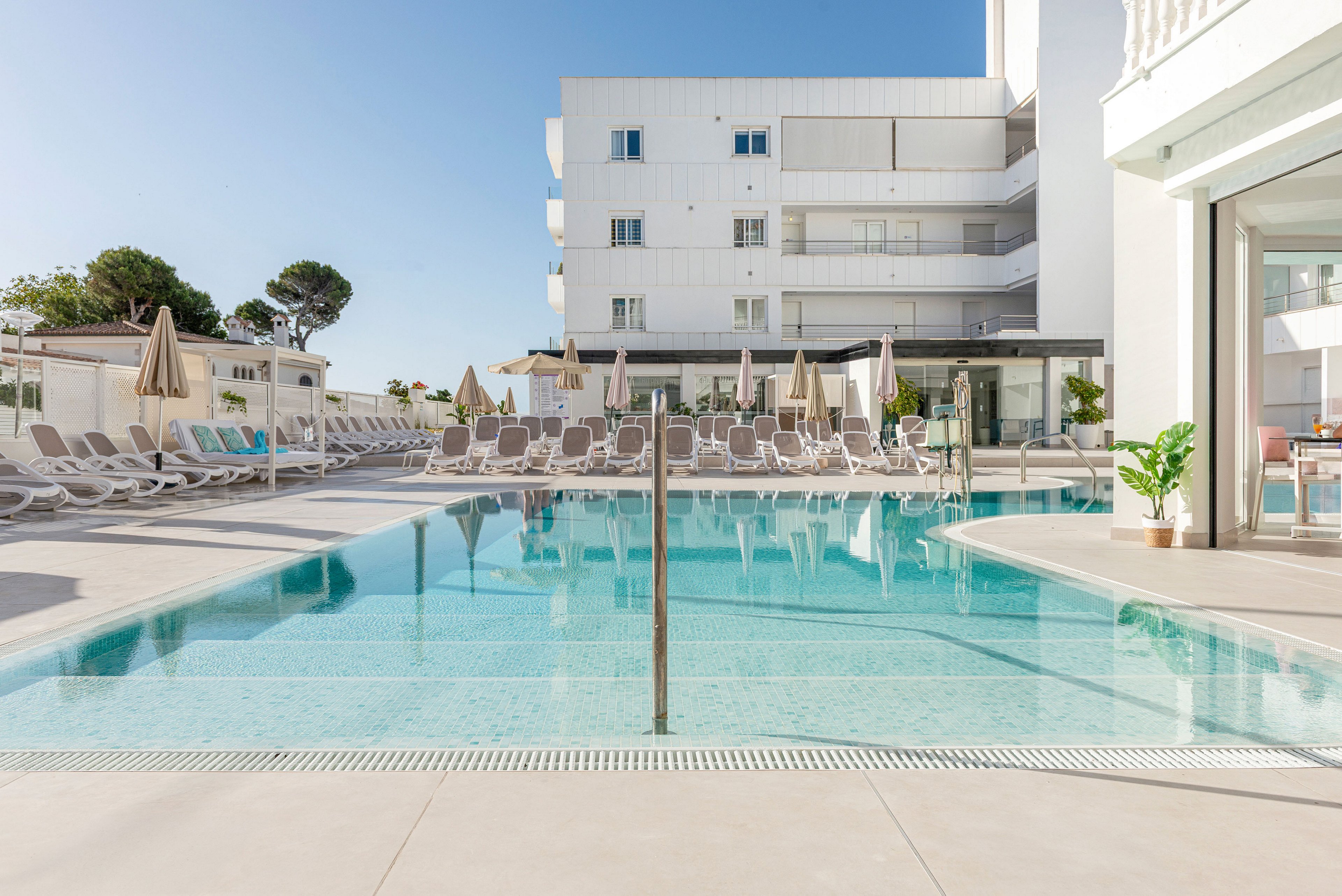 A clear blue swimming pool sits in front of a modern white hotel building. Numerous sun loungers and beige parasols line the poolside terrace, suggesting a relaxing vacation setting. The scene is bright and minimal, with clean architectural lines and soft neutral tones. A few green potted plants add a touch of nature to the contemporary resort environment.