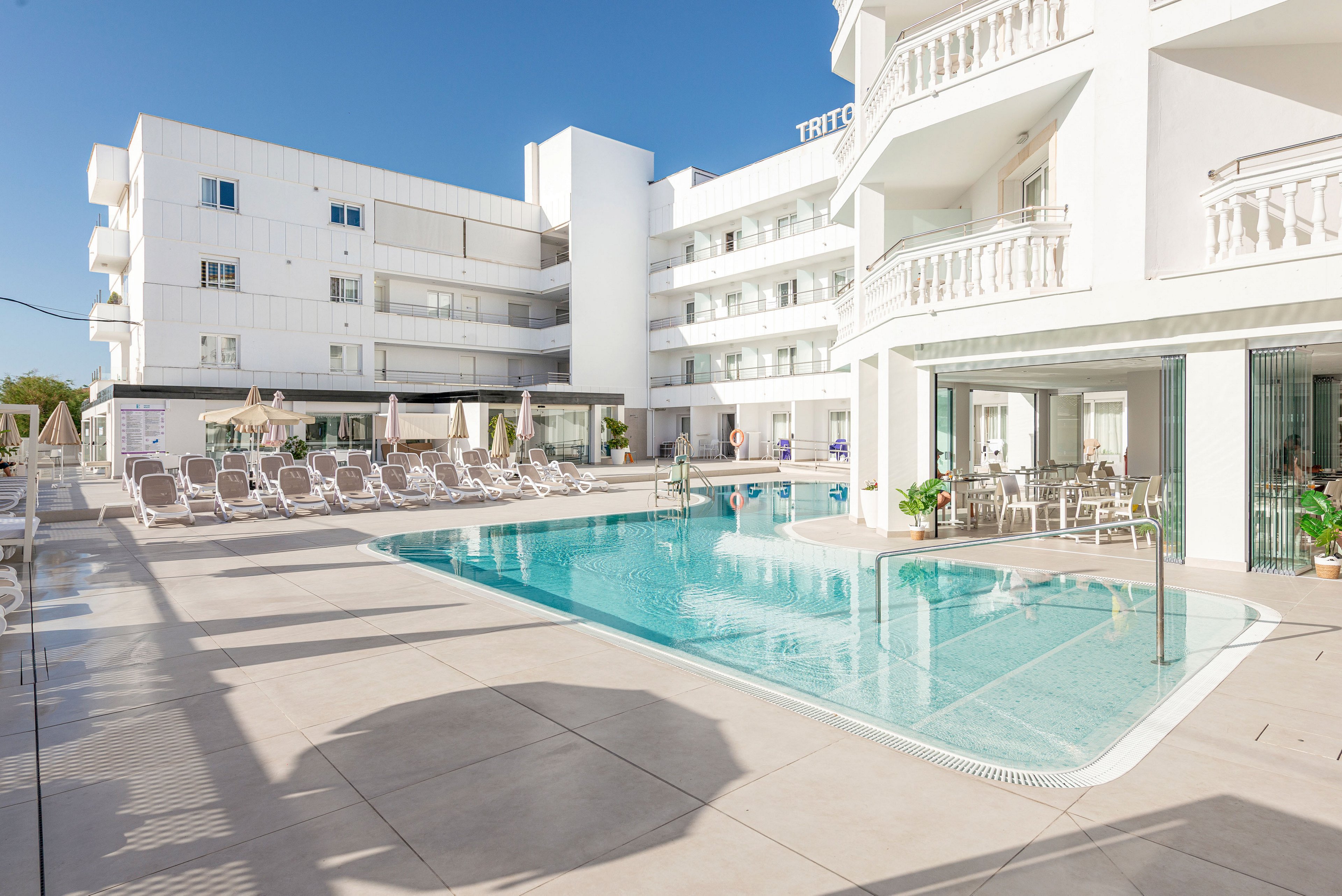 Bright white hotel buildings surround a clear blue outdoor swimming pool in a sunny courtyard. Numerous sun loungers and umbrellas line the poolside terrace, suggesting a relaxing vacation setting. The architecture features balconies and railings, with reflections of the structure visible in the calm water. A few guests are seen near the pool area, enjoying the warm, clear weather under a blue sky.