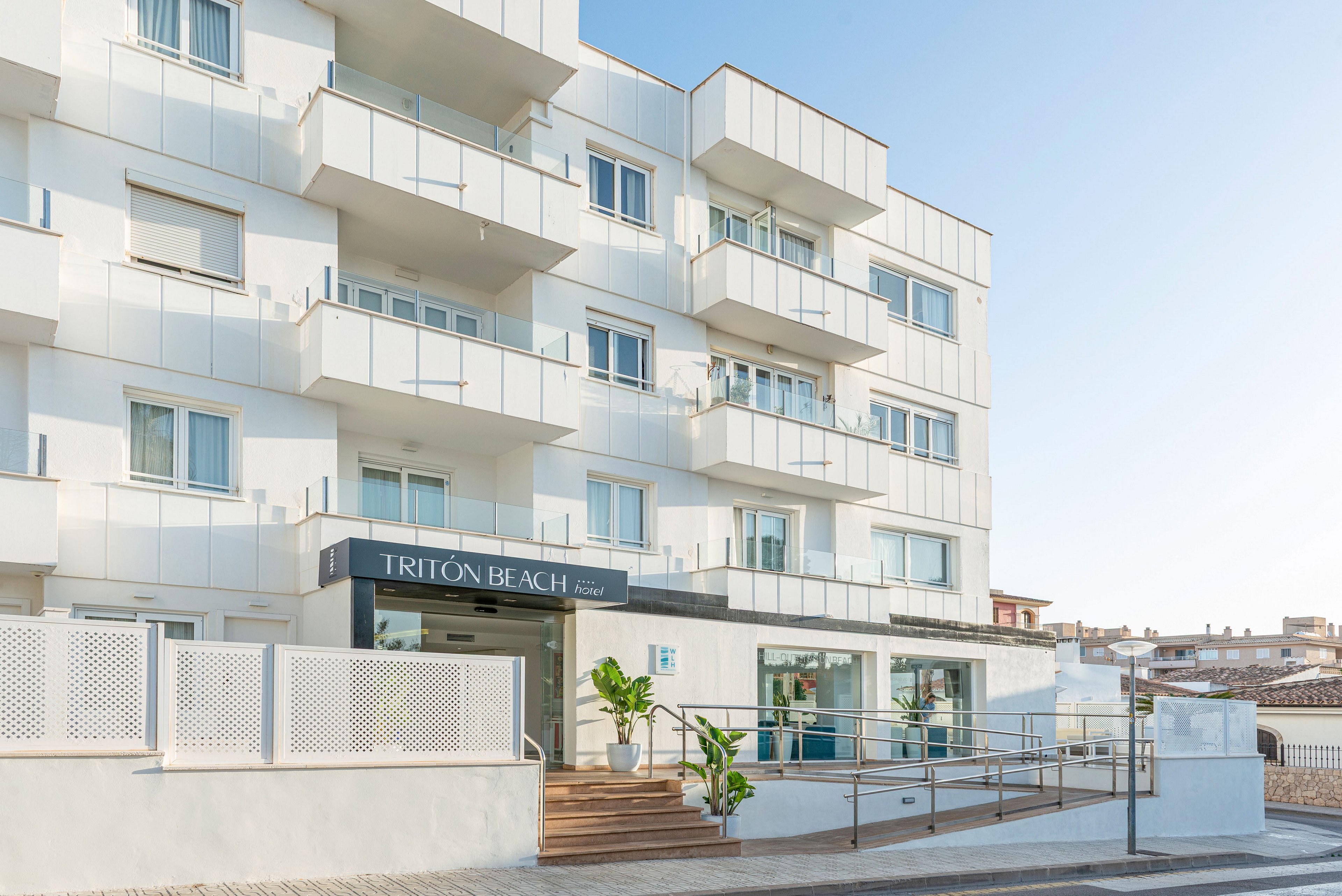 Contemporary white multi-story building houses the Tritón Beach hotel, with its entrance clearly marked by a sign above the doorway. The setting appears to be a sunny coastal town, with clean sidewalks and a gently sloping accessibility ramp leading to the lobby. Large windows and balconies line the facade, while potted plants add greenery near the entrance. The overall scene feels bright, minimal, and welcoming under a clear blue sky.