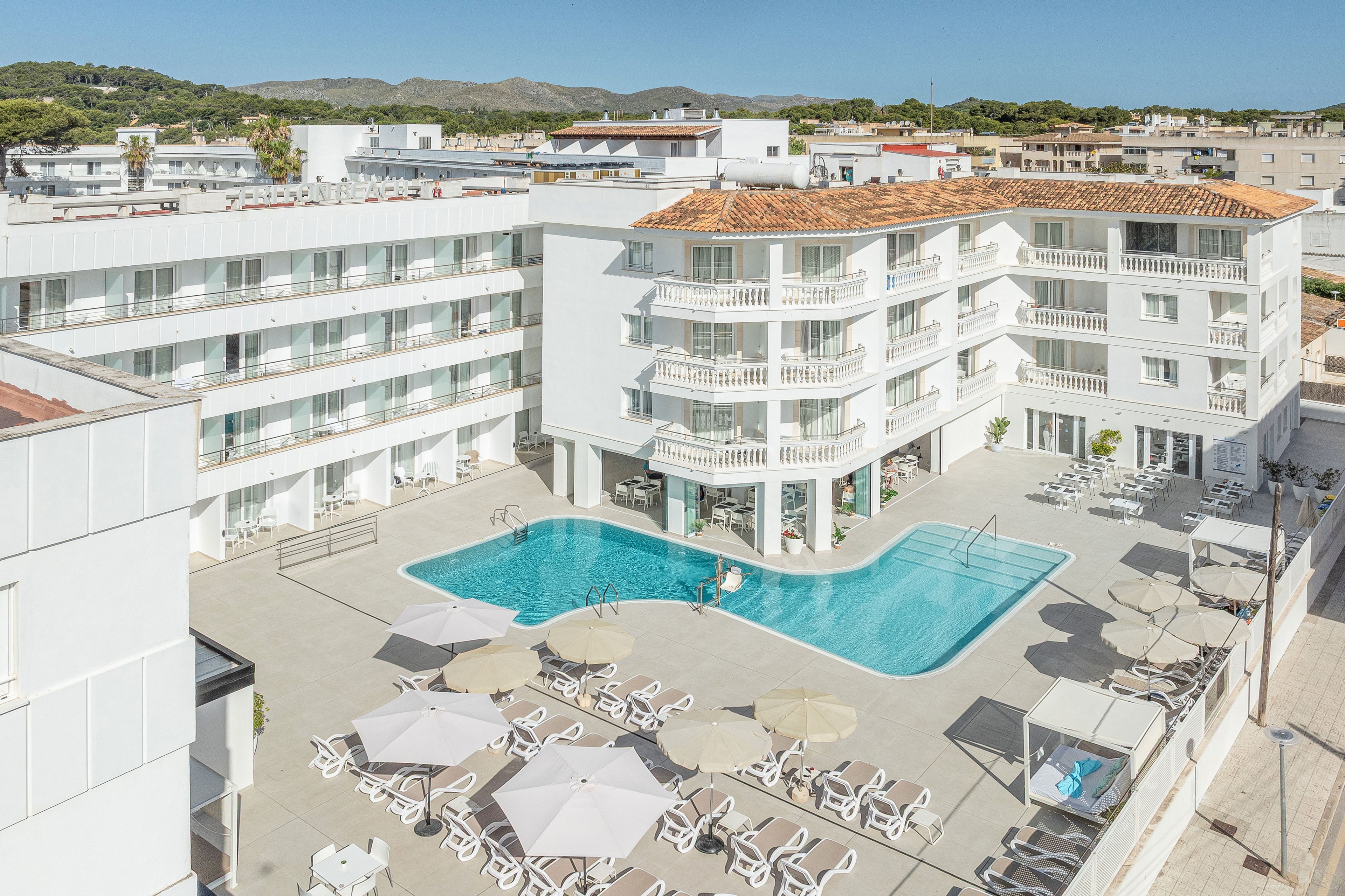 A bright, modern hotel complex surrounds a central outdoor swimming pool. White buildings with multiple balconies frame the sunlit courtyard, creating a clean and minimalist look. Sun loungers and parasols are arranged neatly around the poolside terrace, suggesting a relaxing vacation setting. In the background, low-rise urban structures and distant hills complete the scene under clear daylight.