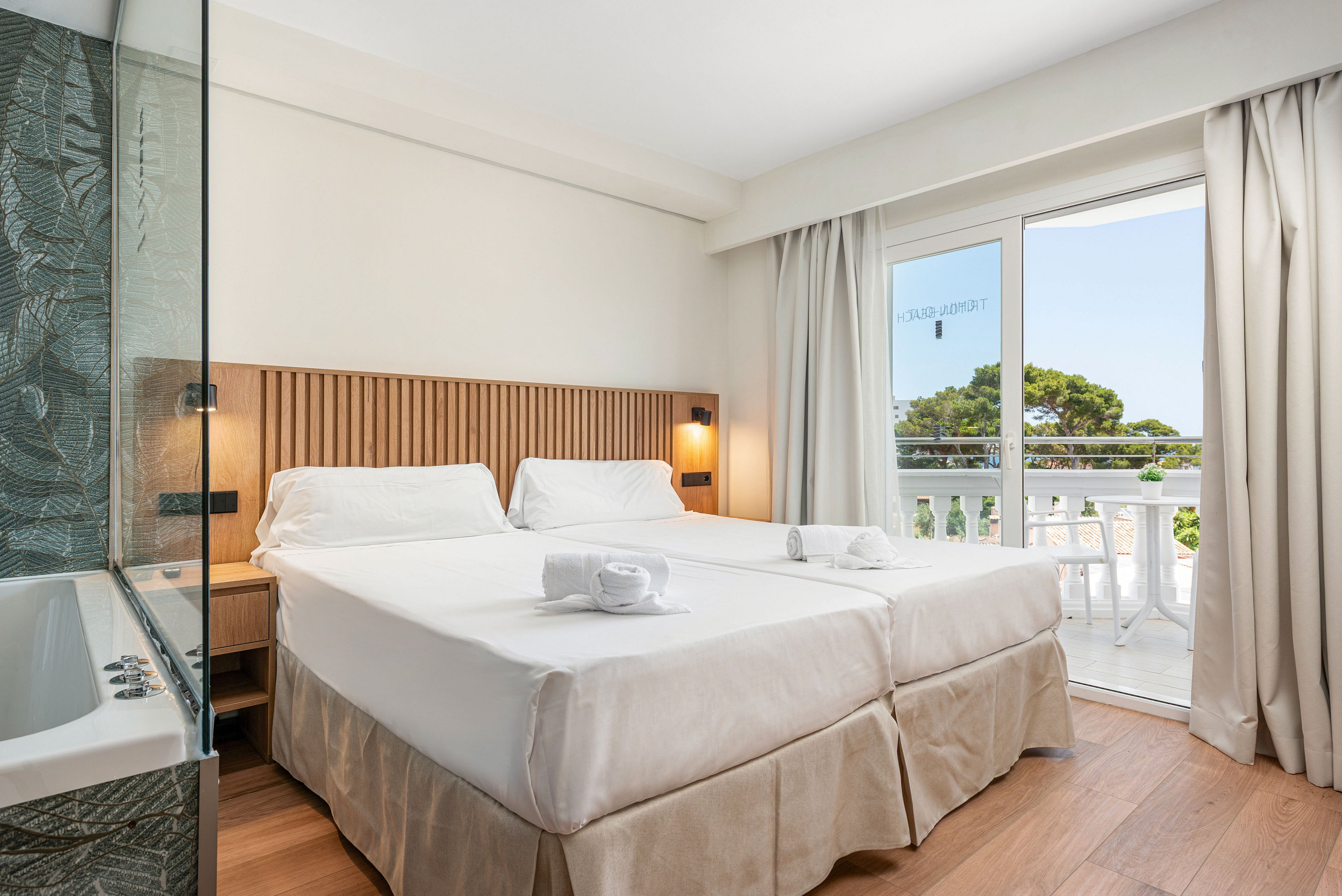 A modern hotel room features two neatly made twin beds with white linens and folded towels placed on top. The space includes a wooden headboard, warm bedside lighting, and a partial view of a bathroom with a bathtub and glass partition. Large sliding doors open to a balcony with white outdoor furniture and a scenic view of trees and blue sky. The overall atmosphere is clean, airy, and inviting, emphasizing natural light and neutral tones.