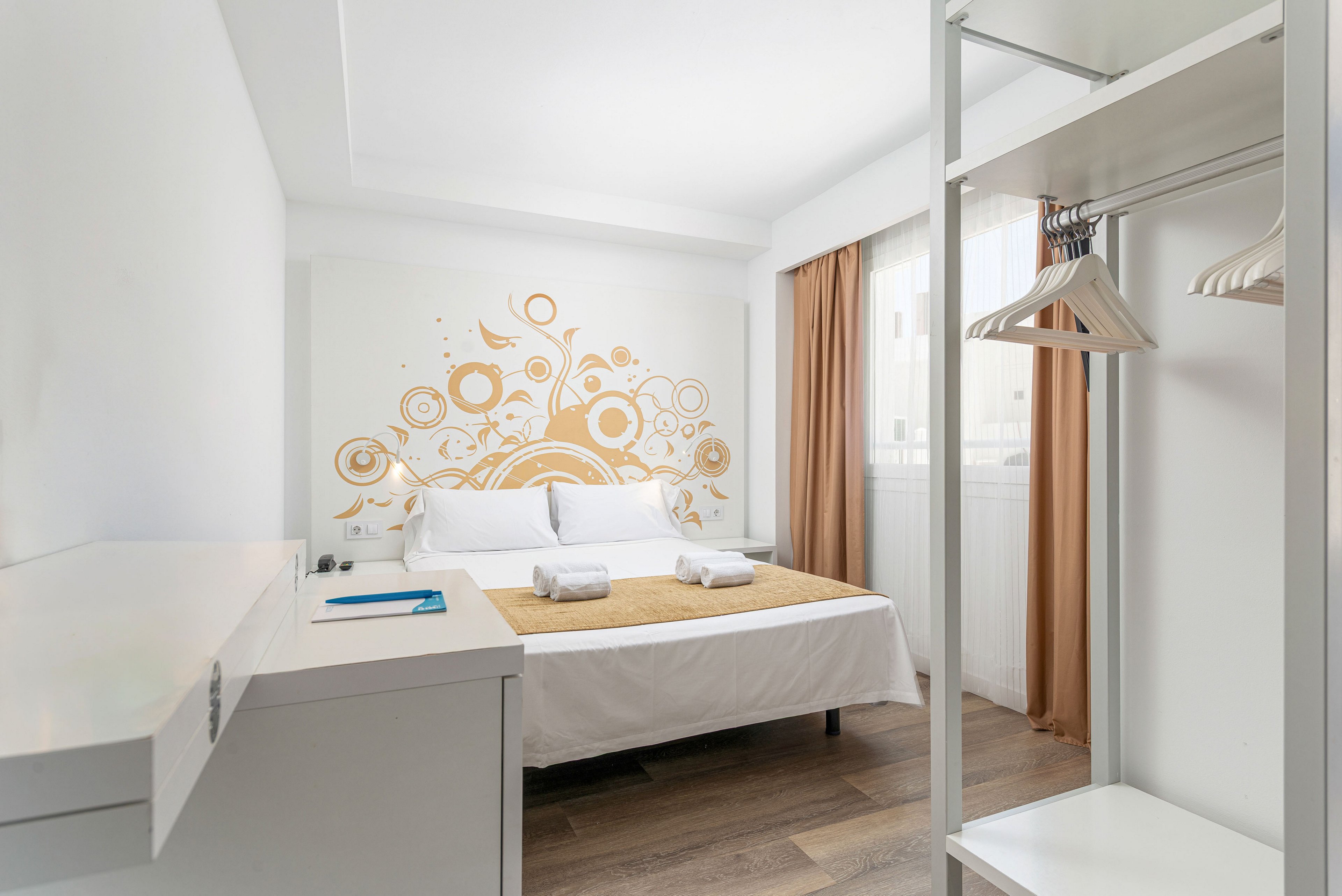 A modern, minimalist bedroom features a double bed with white linens and a golden bed runner. Two neatly folded white towels are placed at the foot of the bed, beneath a decorative wall mural with abstract golden floral patterns. A simple open wardrobe with hangers stands to the right, and a desk with a notebook and pen is positioned to the left. Natural light enters through a window with sheer white curtains and golden drapes, enhancing the clean, airy atmosphere.