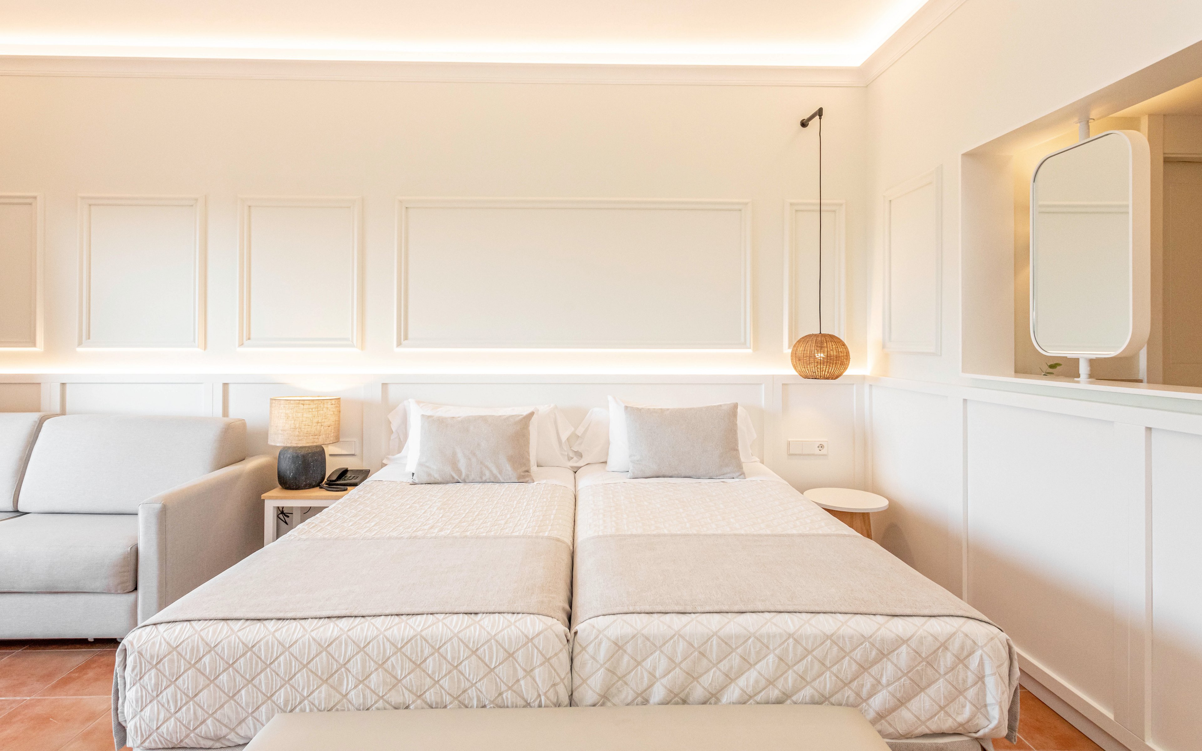 A bright, minimalist bedroom features two single beds pushed together with light quilted bedding. Soft neutral tones dominate the space, complemented by a modern sofa on the left and a padded bench at the foot of the beds. Warm ambient lighting runs along the wall panels and ceiling, with a contemporary pendant lamp and table lamp adding cozy accents. Clean lines, white walls, and subtle decorative details create a calm, modern hotel room atmosphere.