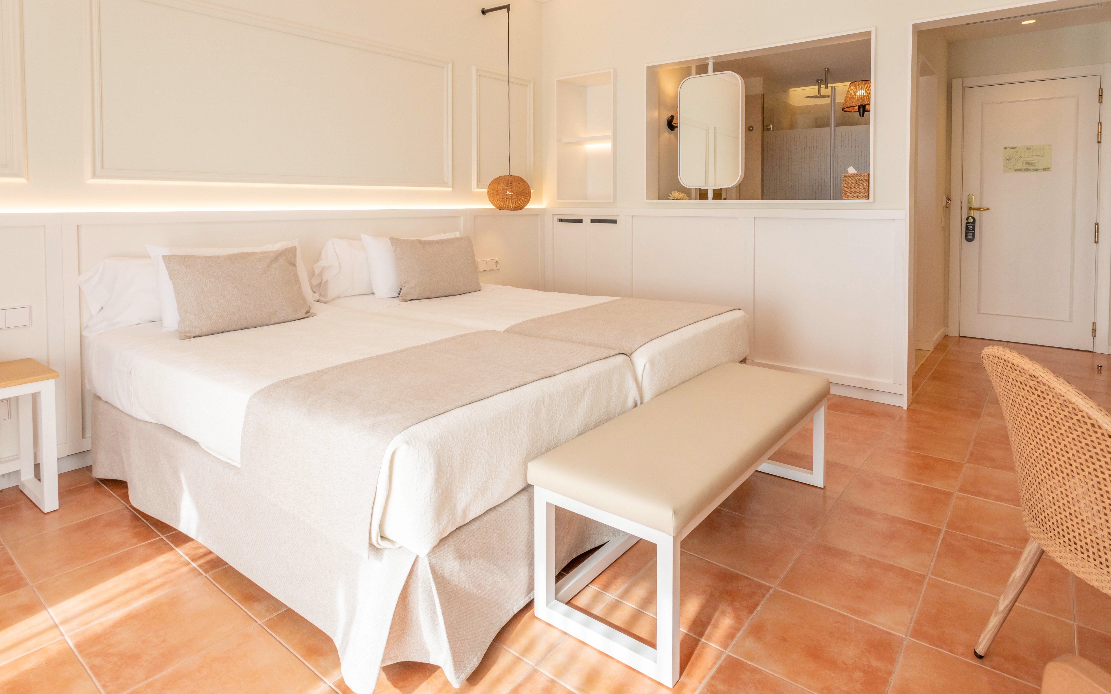A bright, minimalist hotel bedroom features a neatly made double bed with neutral-toned linens and multiple pillows. A padded bench sits at the foot of the bed, matching the light color palette of the room. Warm terracotta floor tiles contrast with the white walls and built-in paneling. A wicker chair, modern lighting, and a partial view of the entry door and wall mirror complete the cozy, contemporary space.