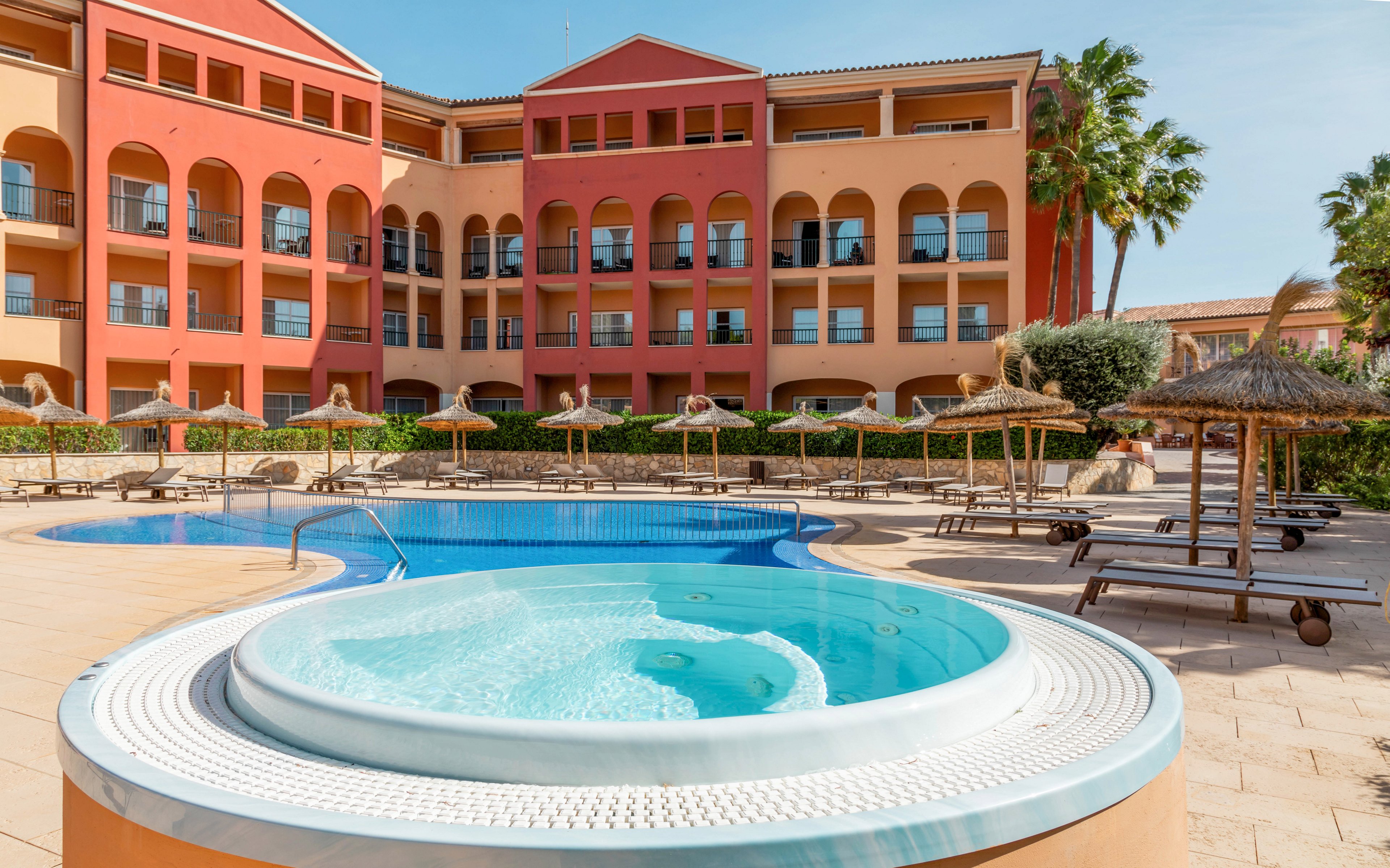 An outdoor jacuzzi sits in the foreground of a sunny resort courtyard. Behind it, a curved swimming pool is surrounded by sun loungers and straw parasols. A multi-story hotel building with warm orange and red facades frames the scene. Tall palm trees and clear blue skies enhance the relaxed vacation atmosphere.