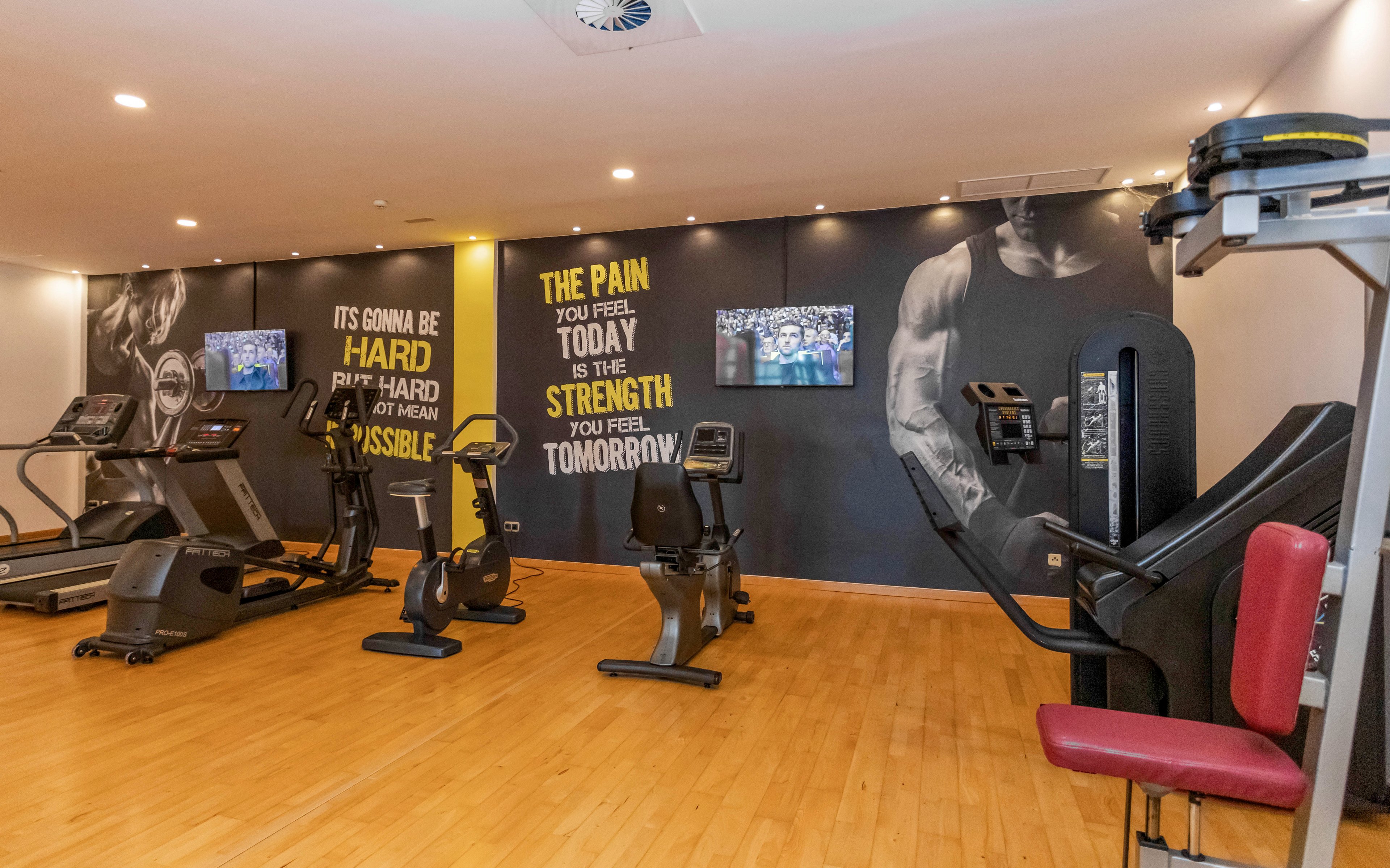 A contemporary gym features several cardio and strength machines arranged on a polished wooden floor. The back wall displays large motivational quotes alongside a black-and-white image of a muscular arm. Two wall-mounted screens show content to entertain people while they work out. Bright lighting and clean finishes create an energetic, professional fitness environment.