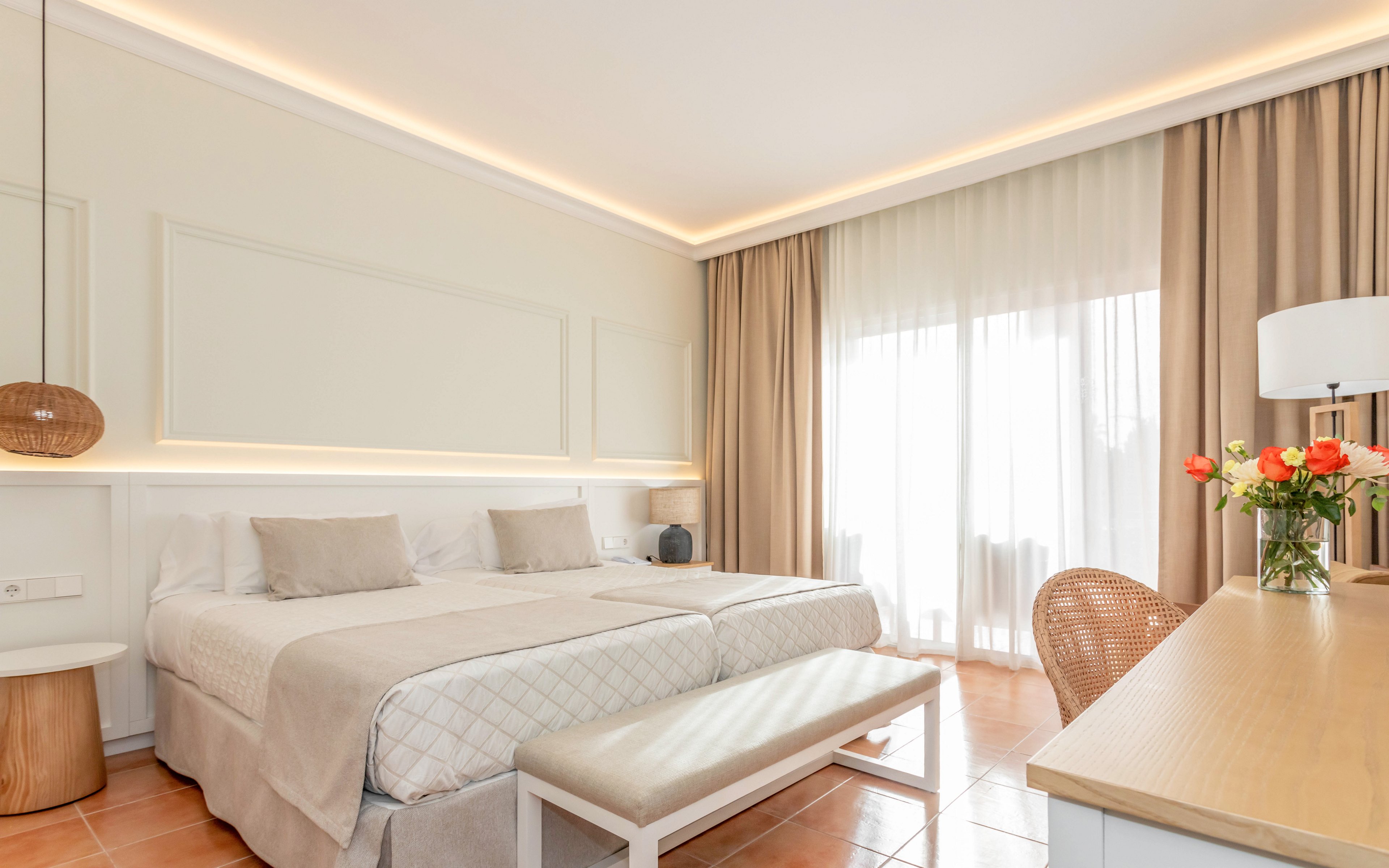 A bright, modern bedroom features two neatly made twin beds with light-colored bedding and cushions. The room is illuminated by soft recessed lighting and natural light streaming through large sheer curtains. A wooden desk with a lamp and a vase of flowers sits opposite the beds, alongside a woven chair. Warm neutral tones and minimalist decor create a calm, inviting atmosphere suitable for a hotel or guest room.