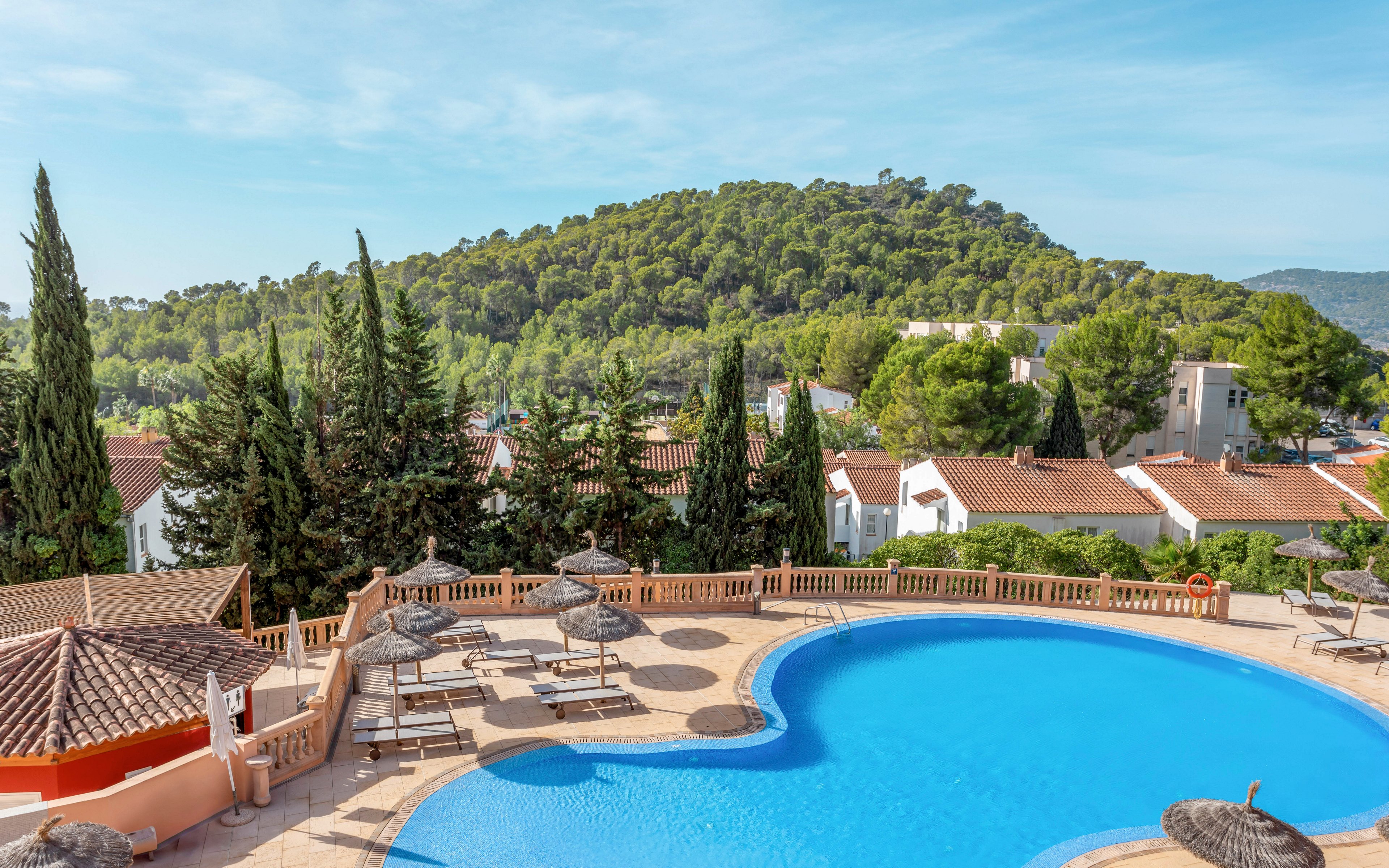 A bright blue resort swimming pool curves in the foreground, surrounded by sun loungers and straw parasols. Terracotta tiles and low walls frame the pool area, giving a Mediterranean feel. Behind the pool, white houses with red roofs sit among tall evergreen trees. A lush, forested hill rises in the background under a clear blue sky, suggesting a peaceful holiday destination.