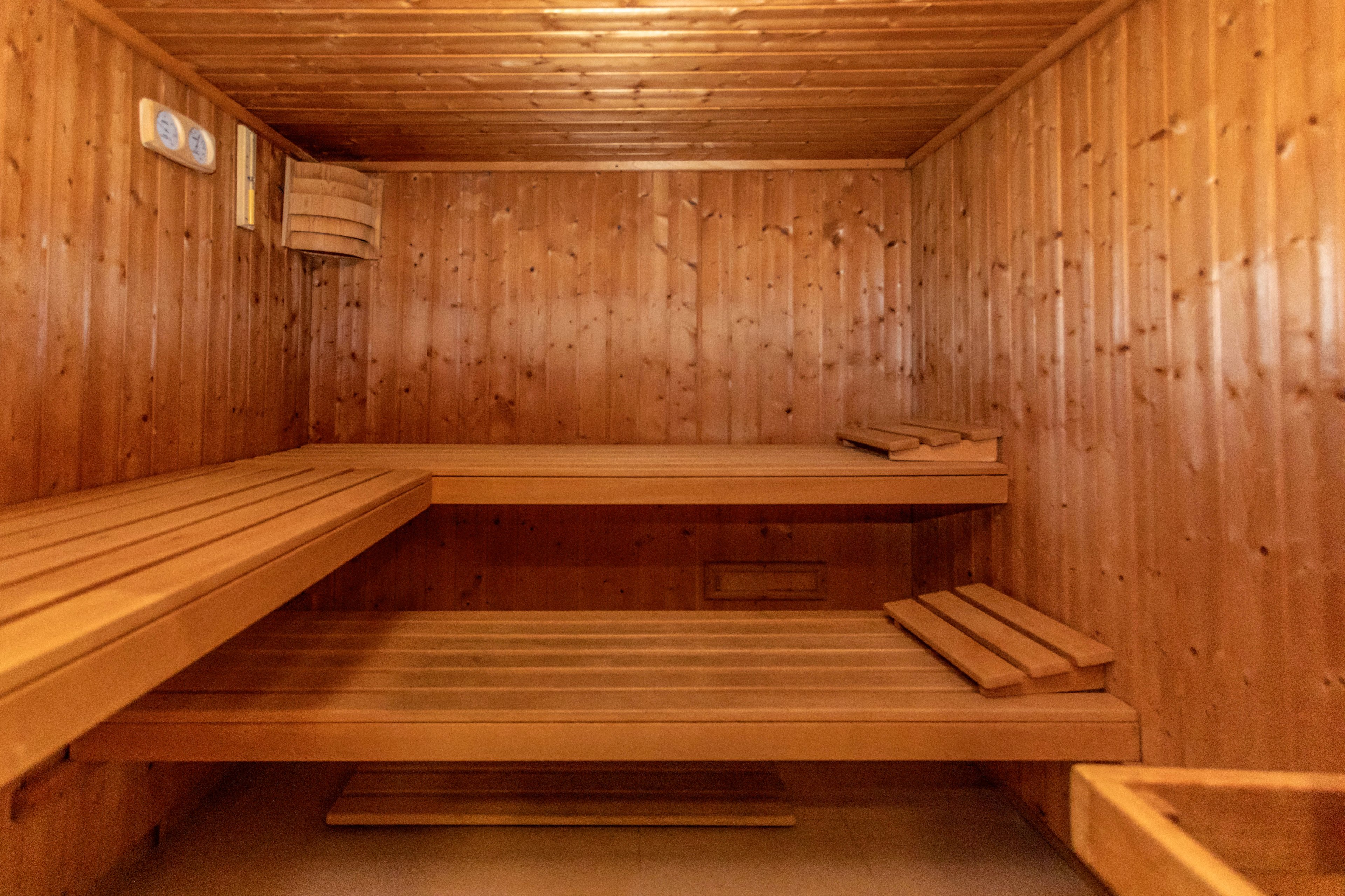 A warm wooden sauna interior is shown with two levels of benches along the walls. The space is empty, highlighting the clean lines and natural wood grain. A wooden light fixture and ventilation elements are visible on the wall and ceiling. The overall scene suggests a calm, relaxing environment ready for use.