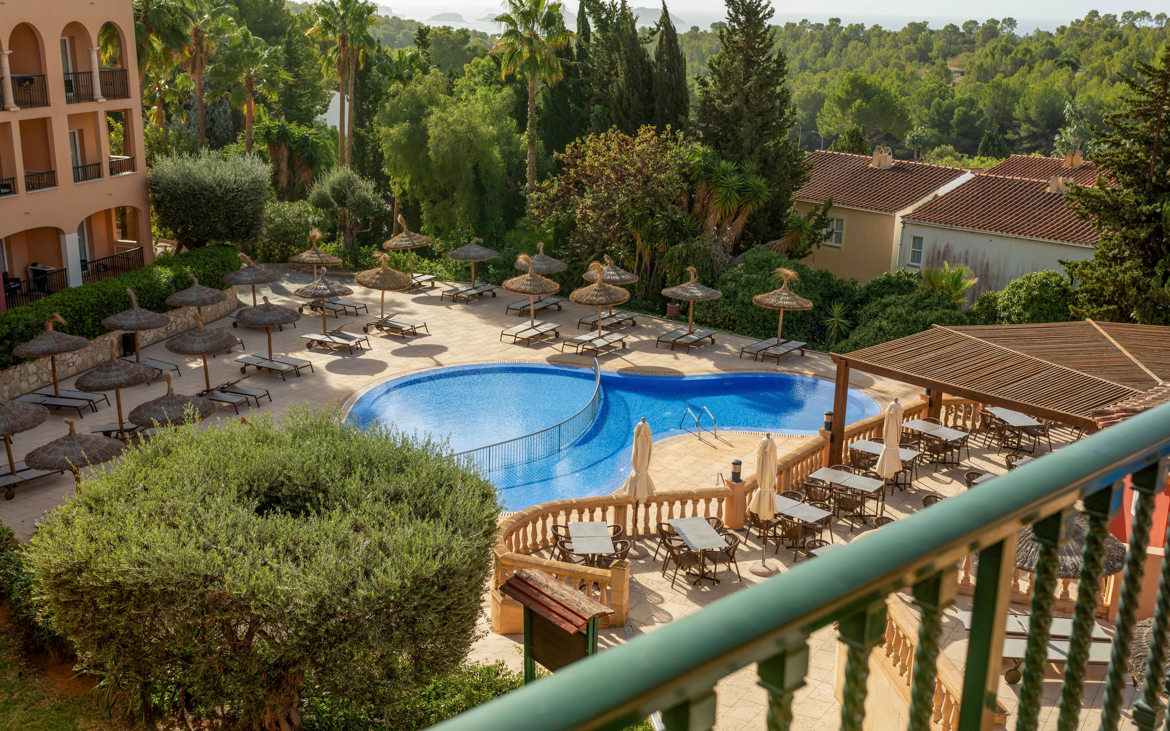 An outdoor resort pool area is viewed from an upper balcony. A curved blue swimming pool is surrounded by numerous sun loungers and straw parasols on a tiled terrace. Green trees, shrubs, and palm trees frame the scene, with residential-style buildings in the background. Warm sunlight and clear weather create a relaxed vacation atmosphere.