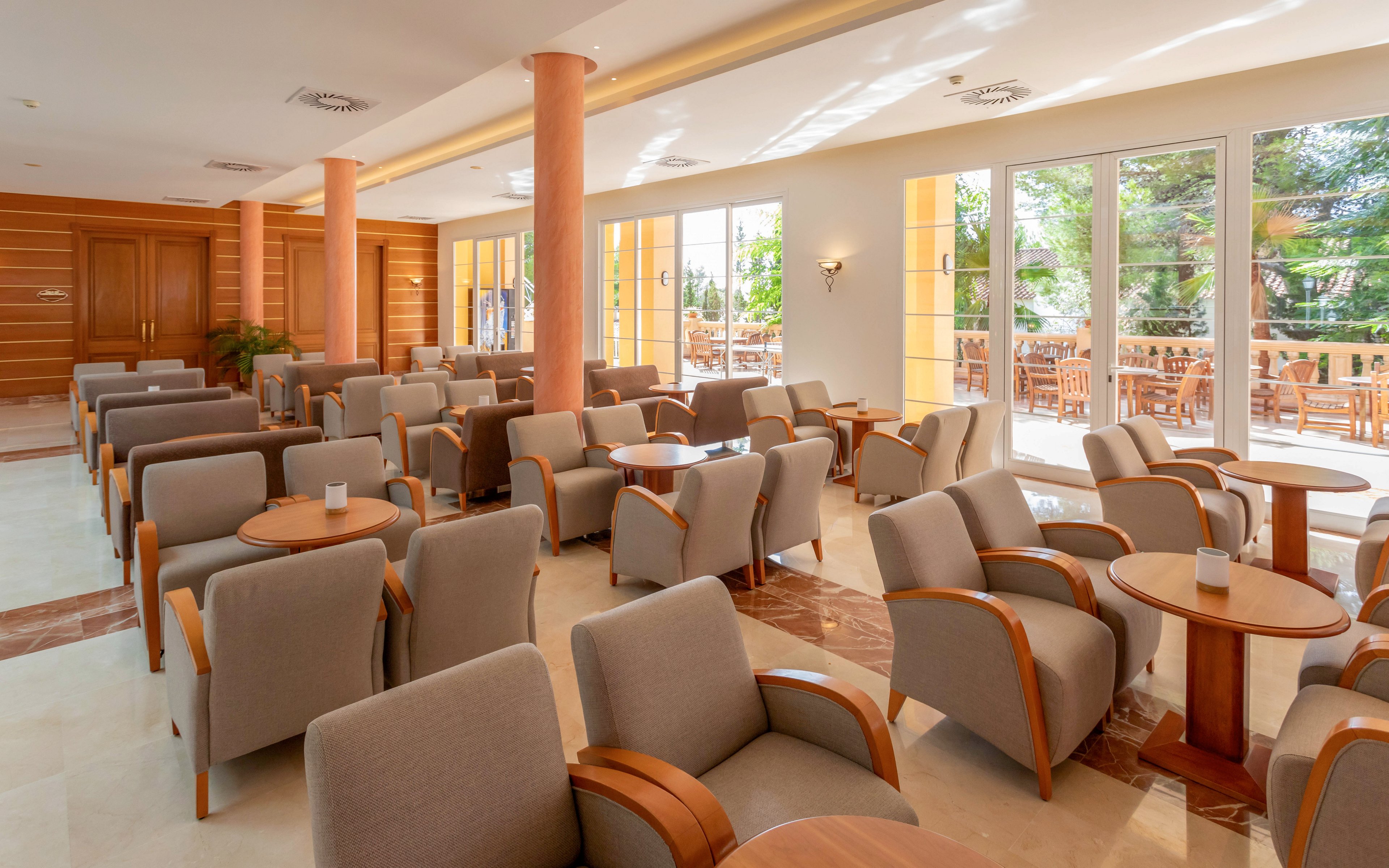 A spacious hotel lounge is filled with rows of beige armchairs and round wooden tables. Large floor-to-ceiling windows and glass doors let in abundant natural light and reveal an outdoor seating area with more tables and chairs. Warm tones from the wooden wall panels, columns, and furniture create a welcoming atmosphere. The room appears clean, quiet, and ready for guests to relax or socialize.
