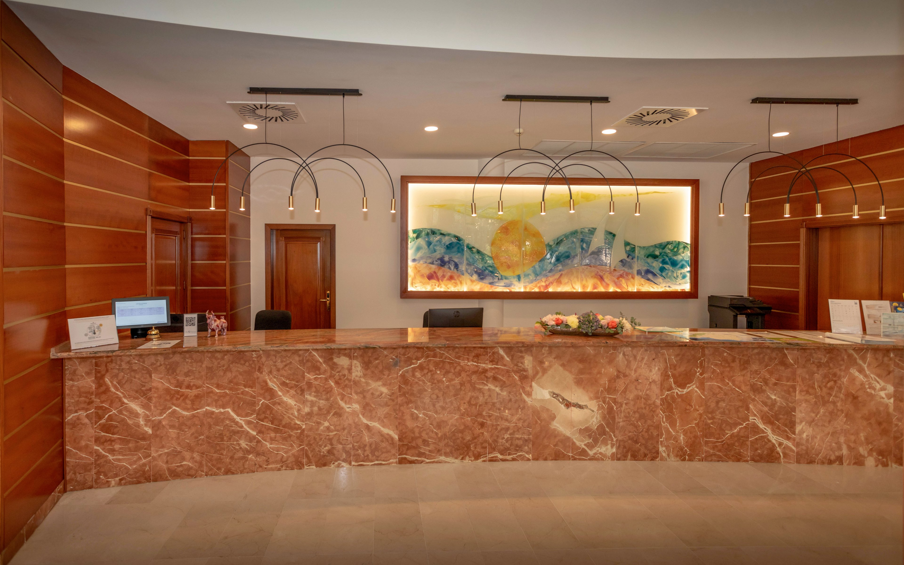 A spacious hotel reception area features a long marble front desk in warm brown tones. Behind the counter, wooden wall panels frame two doors and a large illuminated artwork of a sun over abstract waves. Minimalist pendant lights hang from the ceiling, casting a soft glow over the counter. A computer monitor, documents, and a bouquet of flowers sit neatly on the desk, suggesting a welcoming check-in space. The overall atmosphere is clean, contemporary, and professional.