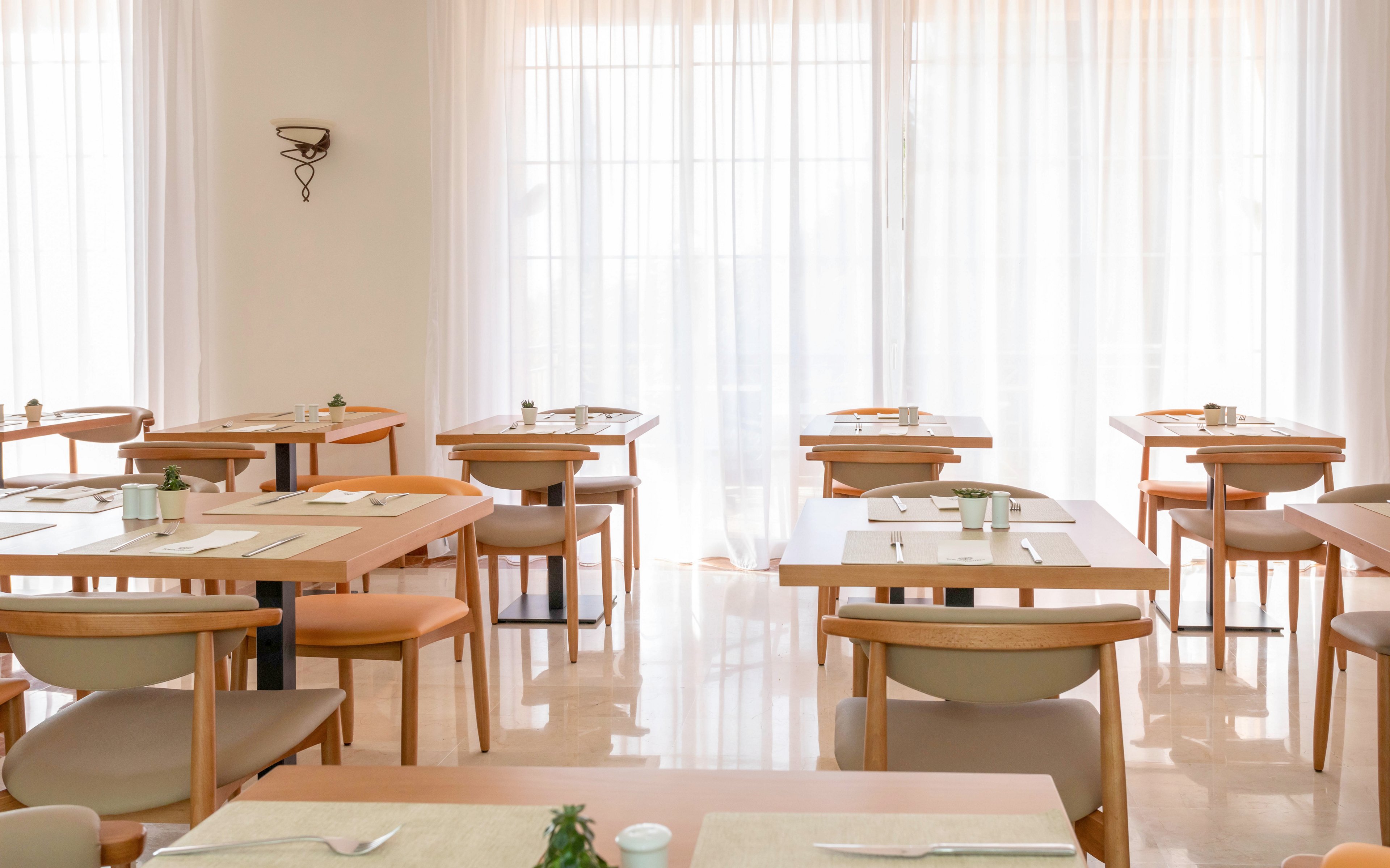 A neatly arranged restaurant dining room features light wooden tables and cushioned chairs. Place settings with napkins and cutlery are prepared on each table, suggesting readiness for guests. Soft daylight filters through sheer white curtains covering large windows, creating an airy atmosphere. The overall scene feels calm, clean, and minimalist with neutral tones and simple decor.