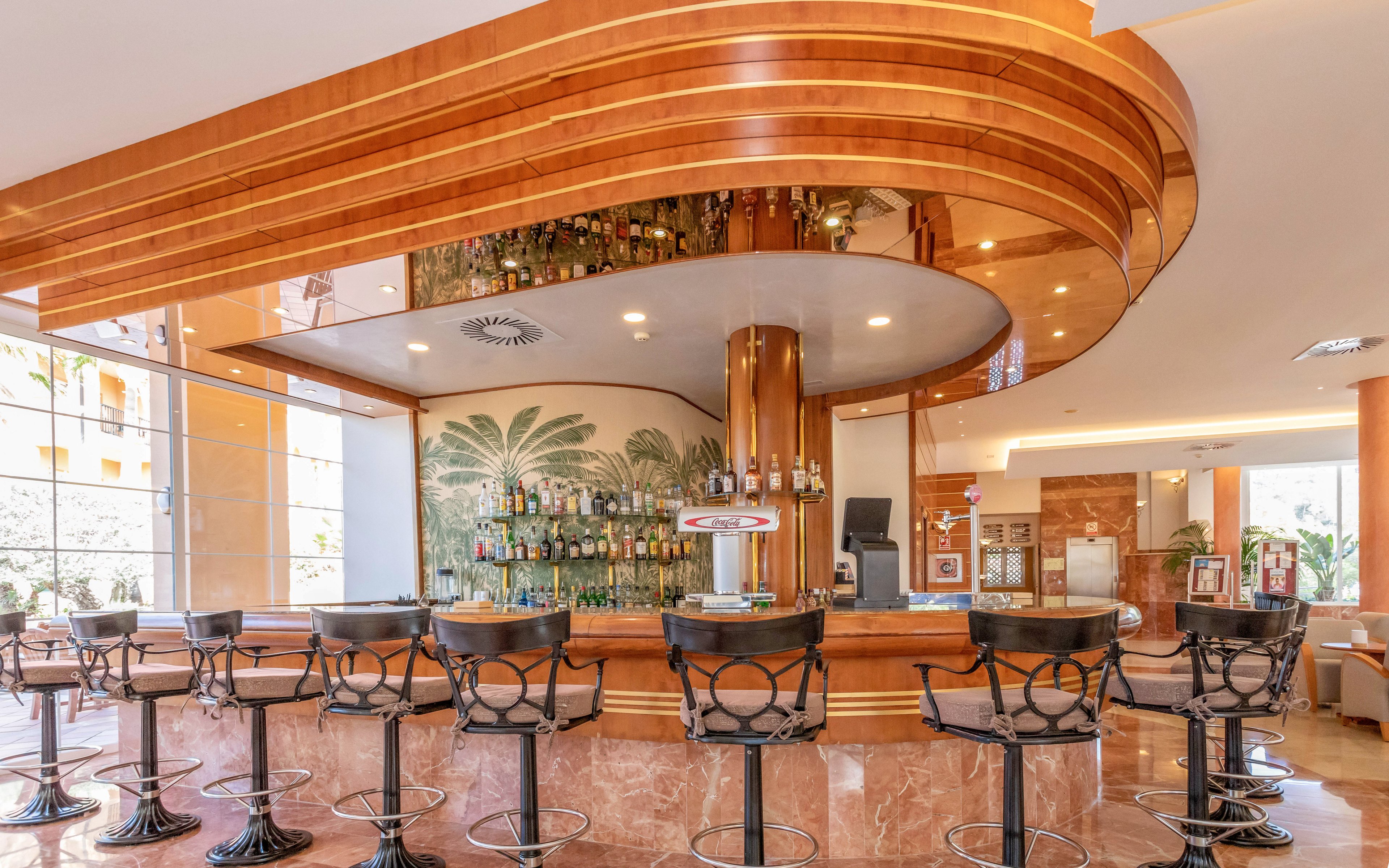 A contemporary hotel bar features a curved wooden counter with high metal bar stools neatly arranged along it. The back wall displays a wide selection of bottles in front of a tropical palm leaf mural. Warm beige and brown tones dominate the interior, complemented by polished marble surfaces and large windows that let in natural light. The space appears clean, inviting, and ready for guests.