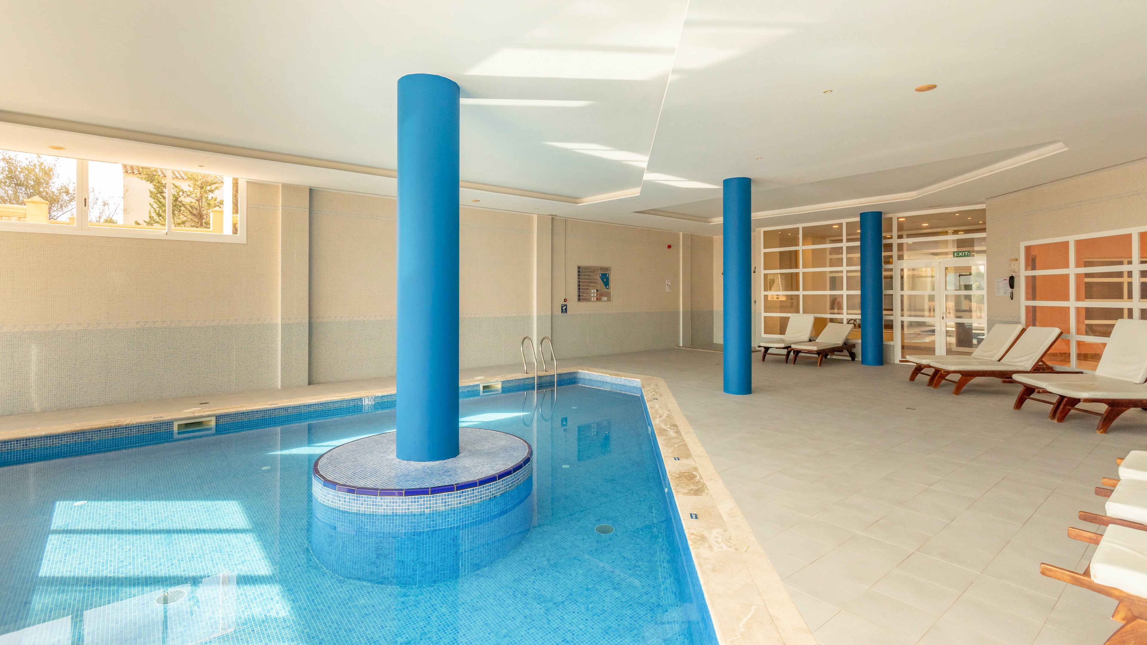 An indoor swimming pool features clear blue water and a central blue column rising from the pool. The space is surrounded by light-colored tiled flooring and several white cushioned lounge chairs arranged along the side. Large windows and glass doors allow natural light to brighten the room, creating a clean and airy atmosphere. The overall setting suggests a relaxing spa or hotel wellness area designed for leisure and comfort.