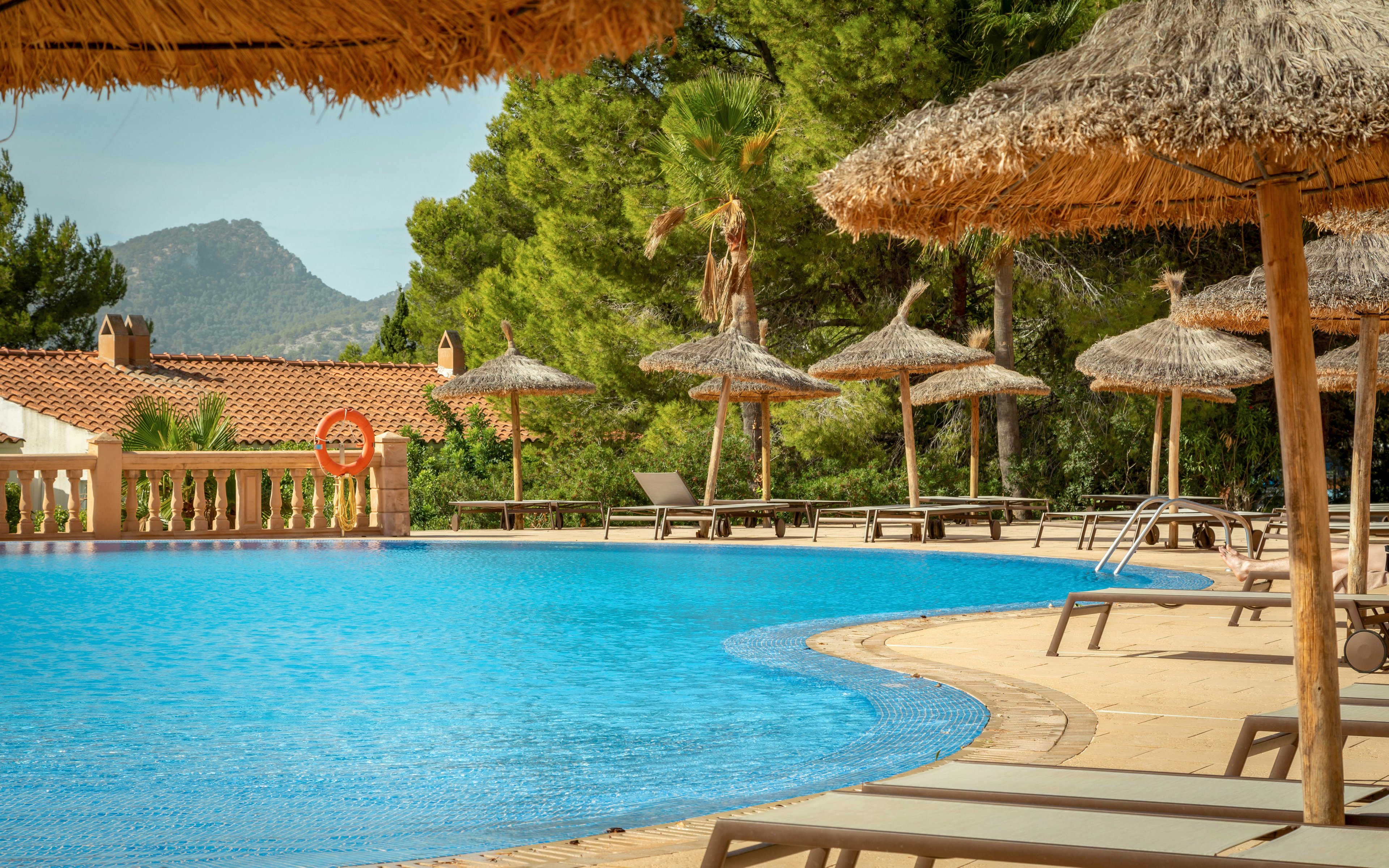 A curved outdoor swimming pool is surrounded by straw parasols and sun loungers at a resort. The clear blue water contrasts with the warm stone pool deck and terracotta-roofed buildings. Pine trees and distant mountains frame the scene, suggesting a Mediterranean holiday destination. The area appears calm and empty, evoking a relaxed vacation atmosphere.
