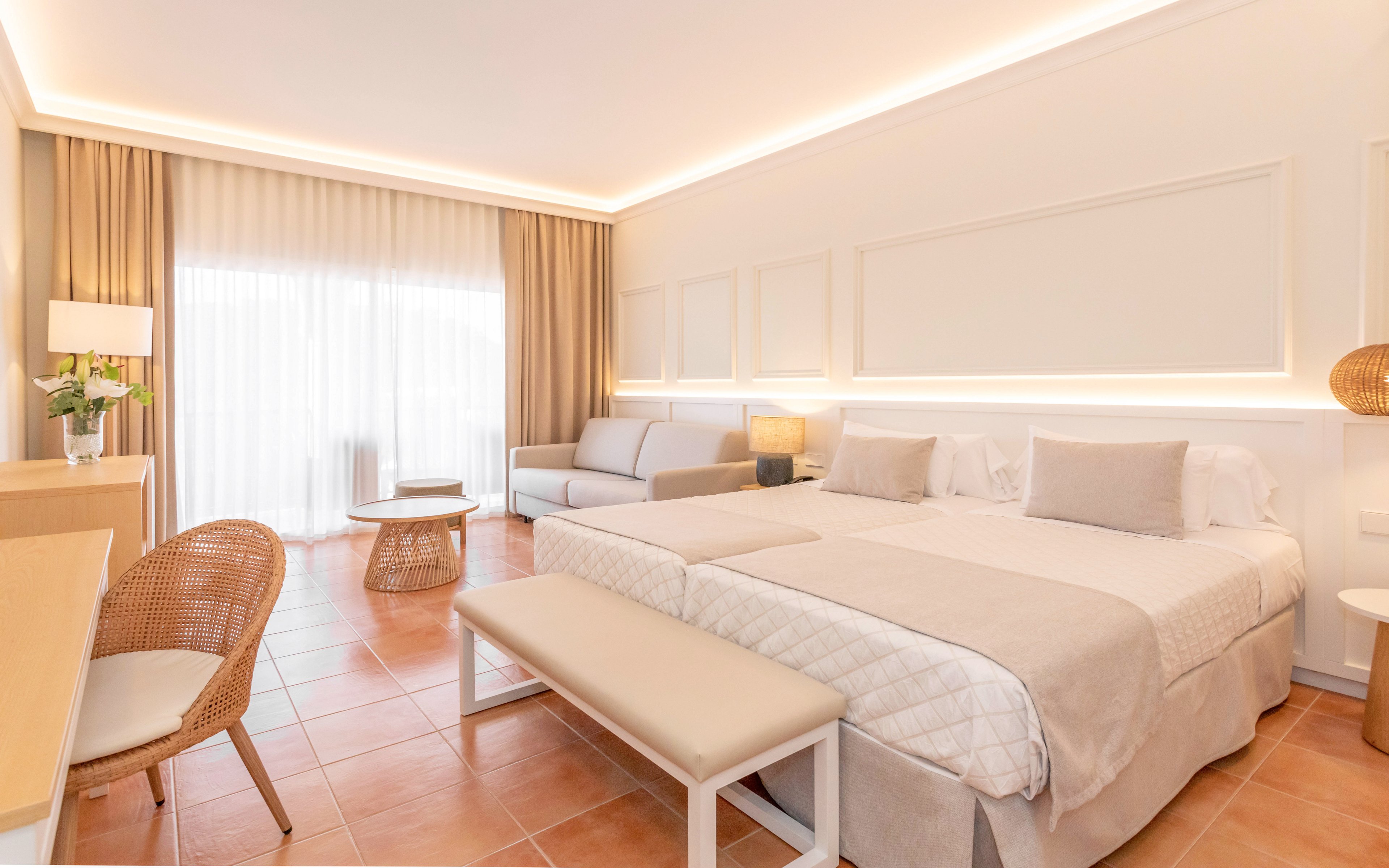 A spacious, modern hotel room features two neatly made twin beds with light-colored bedding and cushions. The room includes a small seating area with a sofa, wicker chair, and side table, all arranged on warm terracotta floor tiles. Large sheer curtains cover a wide window or balcony door, allowing soft natural light to fill the space. Neutral tones, indirect ceiling lighting, and minimalist decor create a calm and inviting atmosphere.