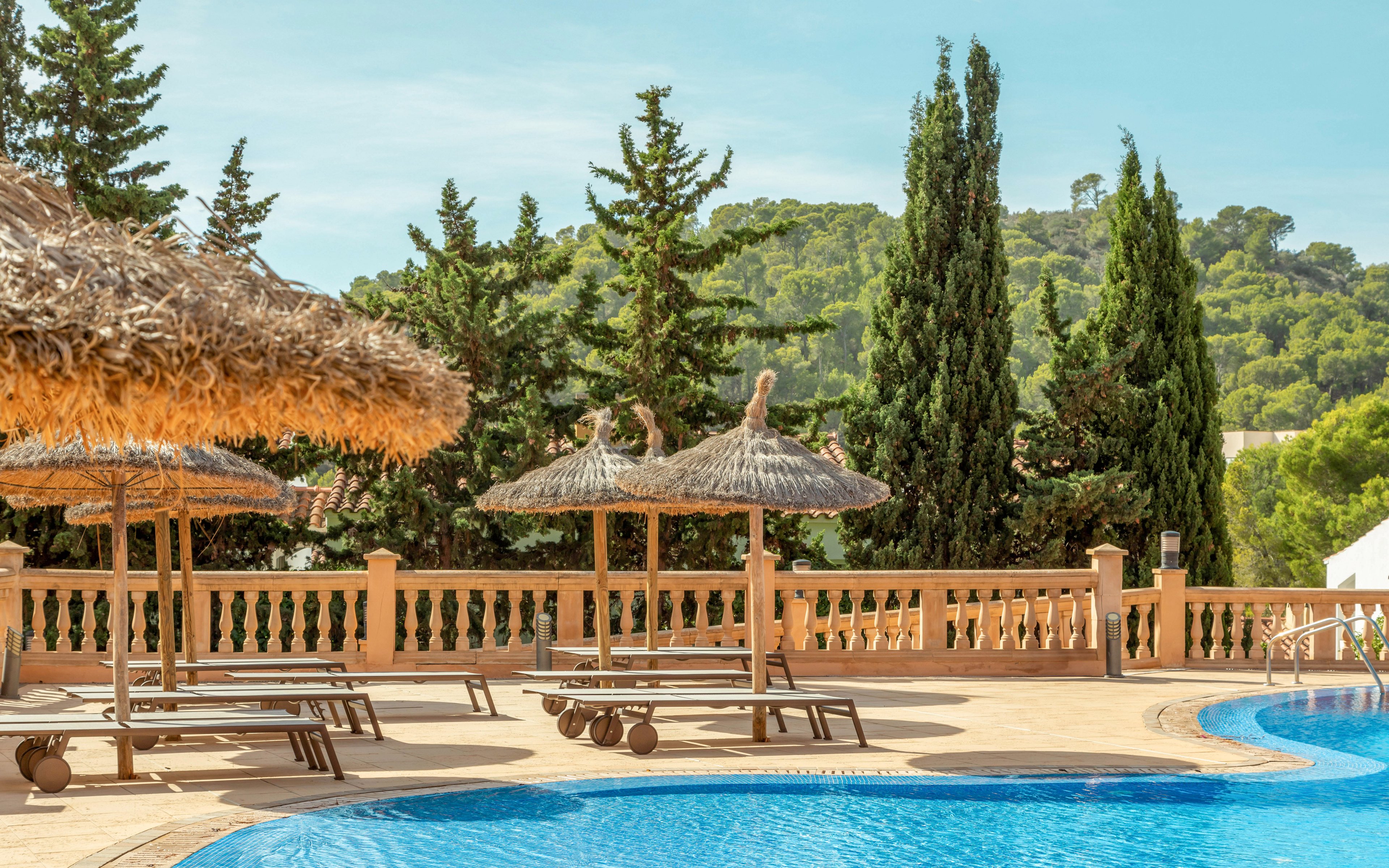 An outdoor swimming pool curves through a sunny resort terrace lined with straw parasols and wooden lounge chairs. A decorative balustrade separates the pool area from a backdrop of tall evergreen trees and rolling green hills. The clear blue water and bright sky create a warm, inviting vacation atmosphere. The scene is calm and empty, emphasizing relaxation and leisure.
