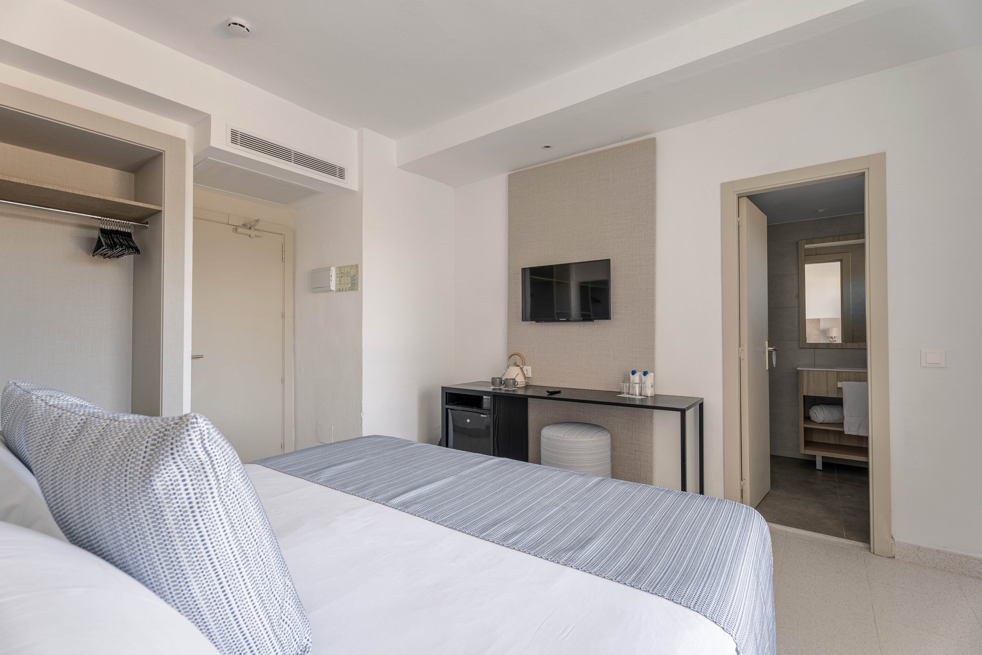 A contemporary hotel bedroom features a neatly made double bed with gray and white linens. The room includes a wall-mounted flat-screen TV above a sleek black desk with water bottles and glasses. An open wardrobe and a closed door are visible on one side, while an open doorway leads to a bright bathroom. The overall design is minimalist with neutral tones and clean lines, creating a calm and orderly atmosphere.