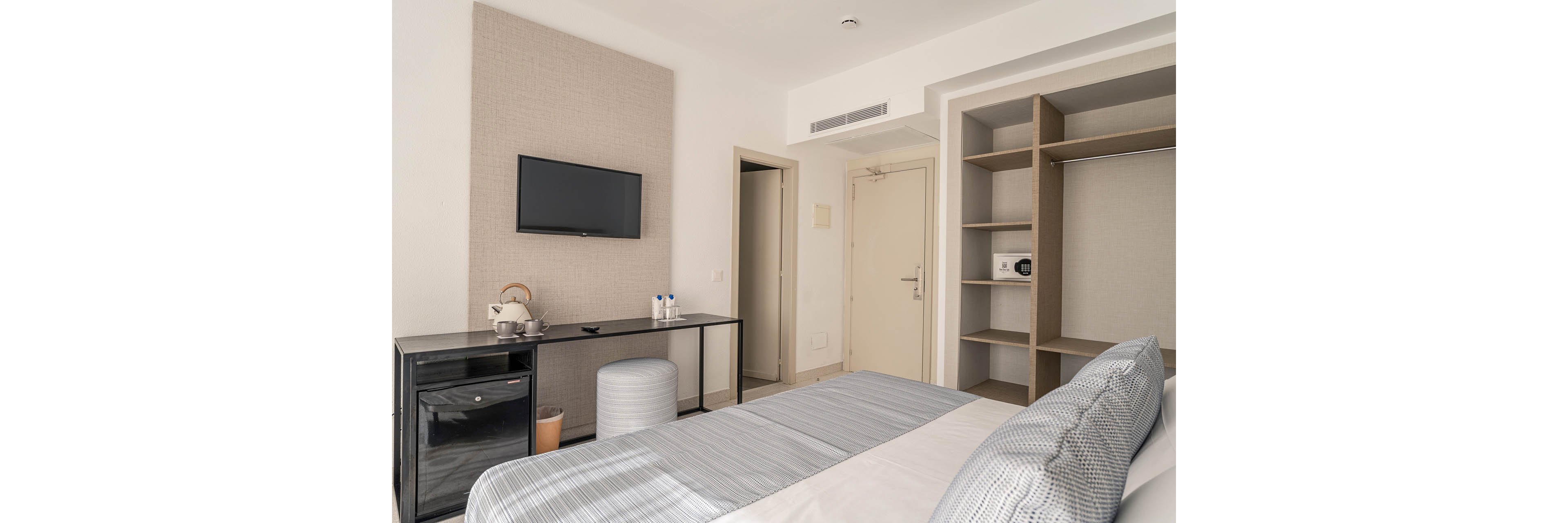 A modern hotel room features a neatly made bed with coordinated gray and white linens. Opposite the bed, a flat-screen television is mounted on the wall above a sleek black console with a kettle and bottled water. Open shelving with a small safe and storage space stands beside the entrance door. The neutral color palette and simple furnishings create a clean, minimalist atmosphere suitable for business or leisure stays.