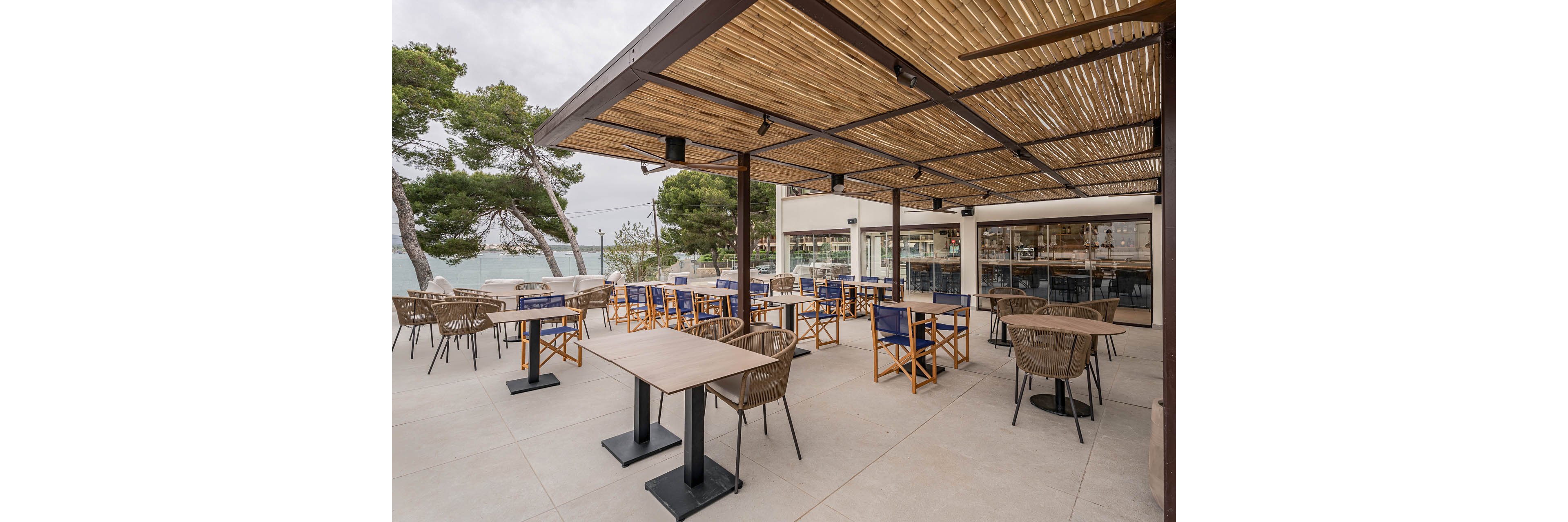 An open-air terrace café features neatly arranged tables and chairs under a slatted wooden pergola. The setting overlooks a coastal area with trees and water visible in the background. Large windows of the adjacent building reflect the outdoor seating area, enhancing the airy, modern atmosphere. The scene appears calm and quiet, with no people present and neutral tones dominating the palette.
