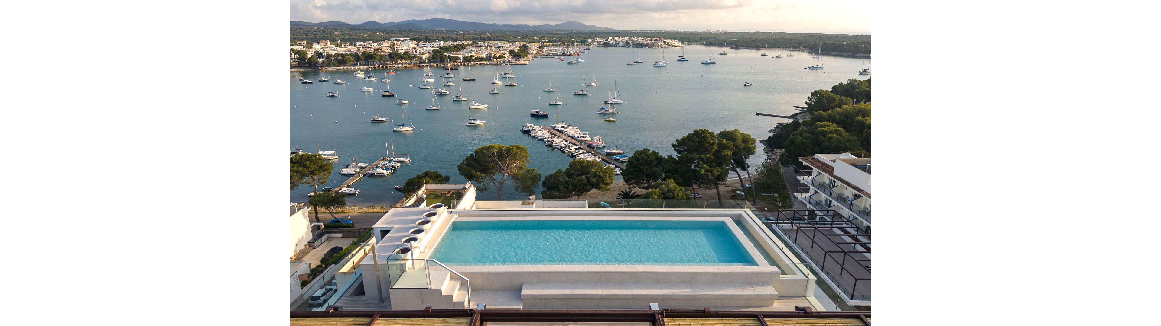 A modern rooftop infinity pool sits above a coastal town, offering a panoramic view of a calm marina filled with anchored boats. Contemporary white architecture and glass railings frame the turquoise water of the pool. Beyond, the shoreline curves around the bay, lined with trees, low-rise buildings, and distant hills under a soft sky. The overall scene suggests a luxurious, tranquil vacation setting by the sea.