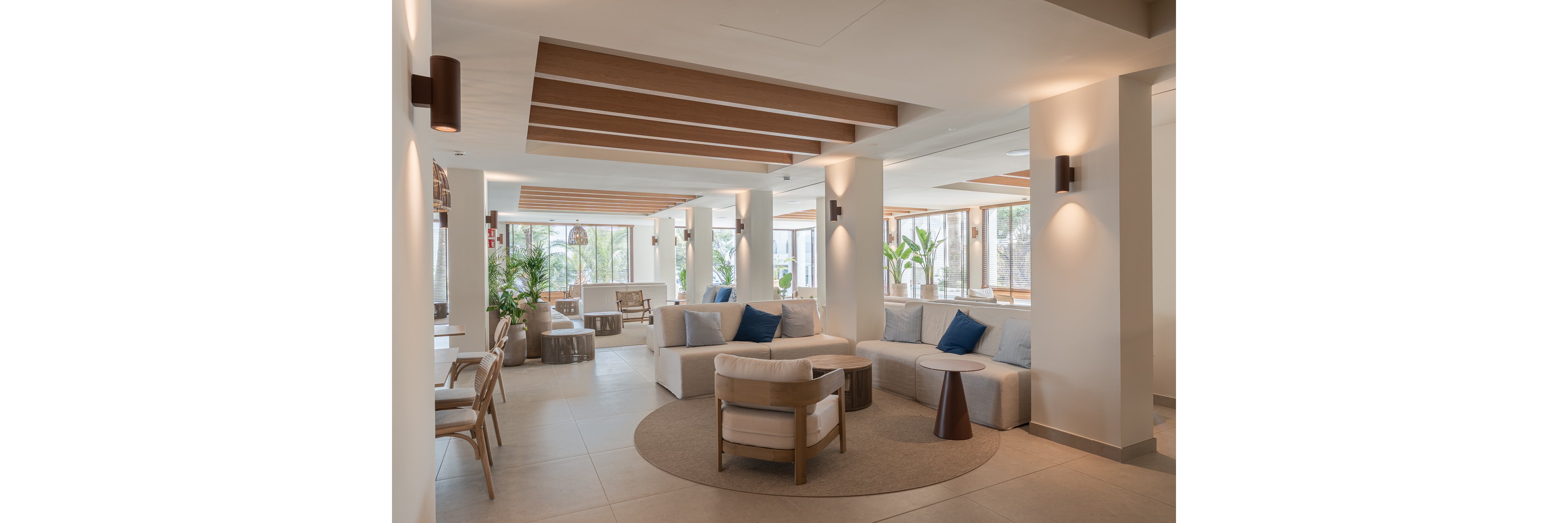 Spacious modern lounge area with neutral-toned sofas and armchairs arranged for relaxed seating. Large windows fill the room with natural light and reveal greenery outside. Wooden ceiling beams and minimalist wall sconces add warm architectural detail. Potted plants, side tables, and soft cushions create a calm, welcoming atmosphere suitable for a hotel lobby or upscale waiting area.