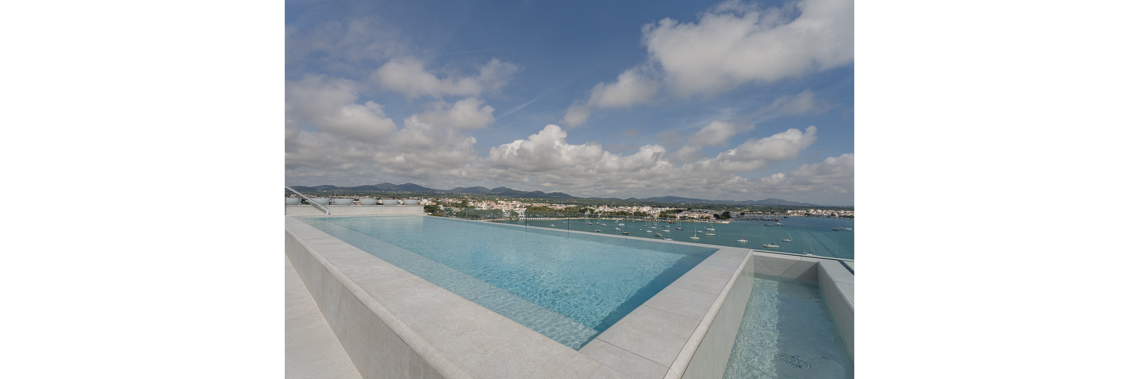 A modern rooftop infinity pool stretches toward a scenic coastal bay. Calm blue water reflects the partly cloudy sky, creating a serene atmosphere. In the distance, a small town with white buildings lines the shoreline, backed by rolling green hills. Boats dot the water, enhancing the relaxed vacation feel of the scene.