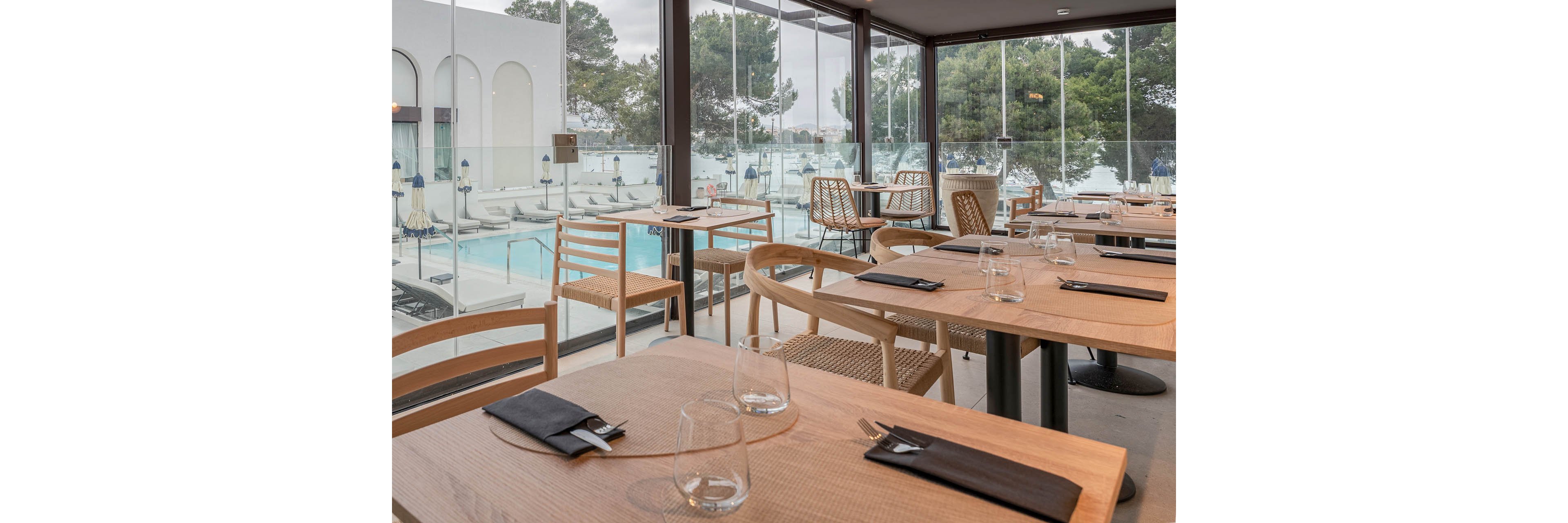 Contemporary wooden tables are set with glasses and black napkins inside a modern restaurant. Floor-to-ceiling glass walls reveal an outdoor swimming pool area with sun loungers and umbrellas. Trees and a calm waterfront can be seen in the background, creating a serene resort atmosphere. The overall scene feels clean, minimal, and ready for guests.