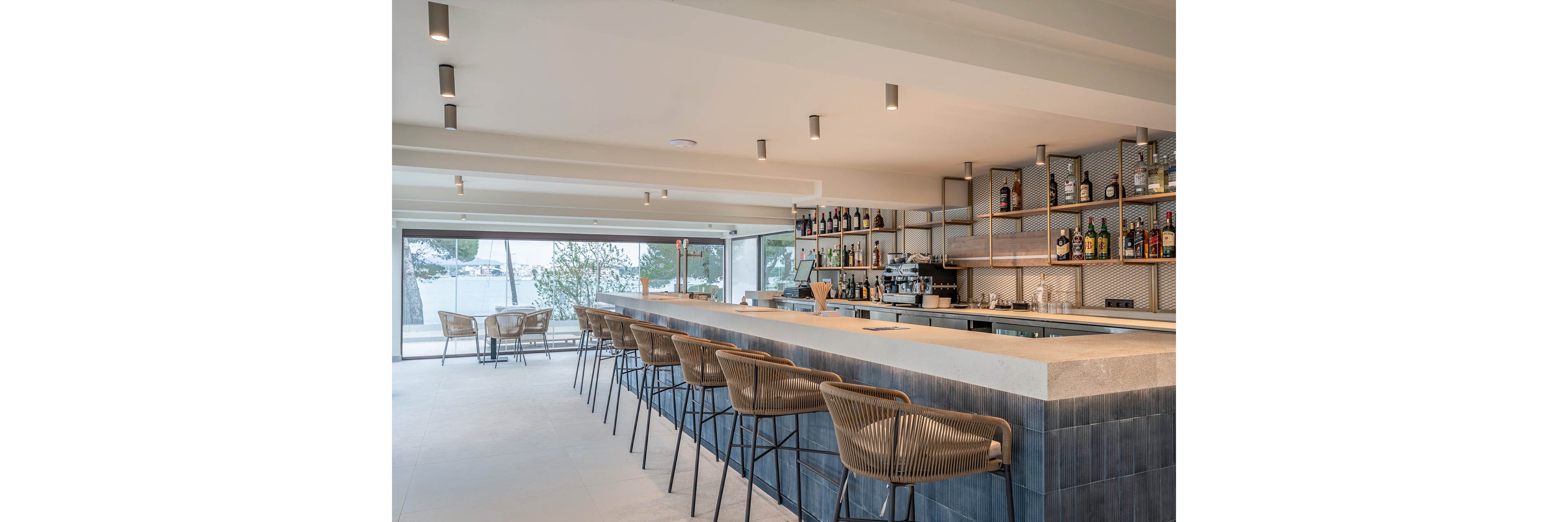 A contemporary bar interior features a long stone counter with high woven stools neatly arranged along its length. Shelving behind the bar displays an assortment of bottles and glassware, with a coffee machine and other bar equipment visible. Large windows open to an outdoor seating area and provide views of trees and a waterfront setting. Neutral tones, clean lines, and recessed ceiling lights create a bright, minimalist atmosphere suited for relaxed socializing.
