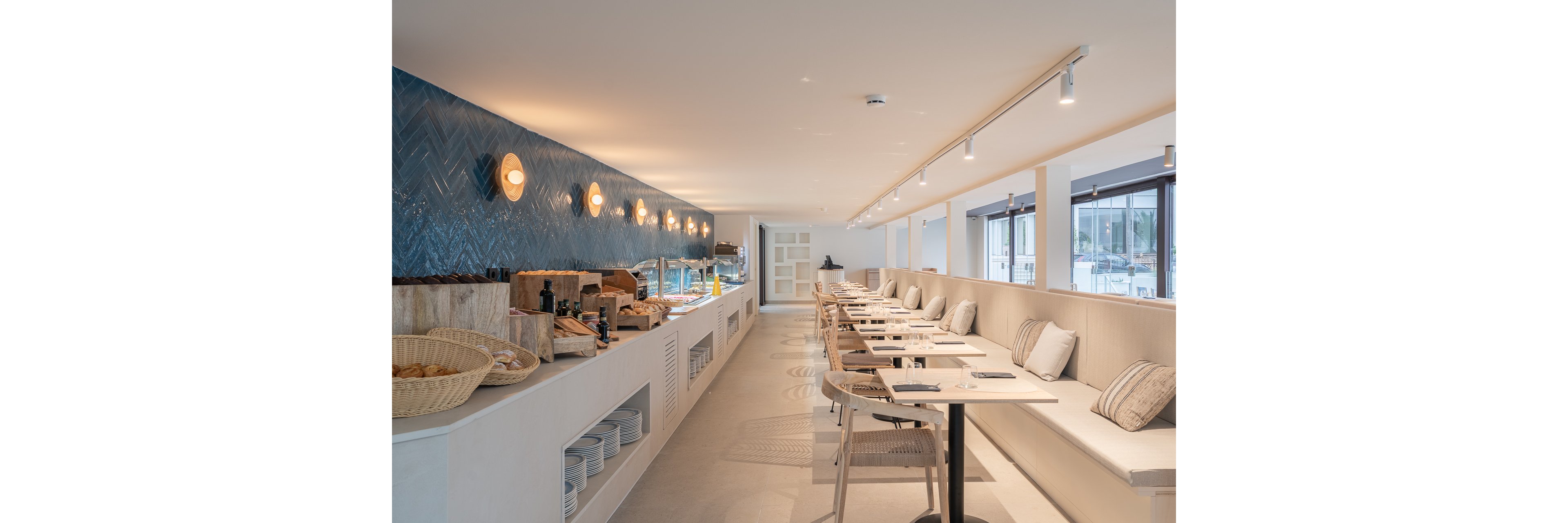 A contemporary restaurant interior features a long buffet counter on the left and built-in bench seating on the right. The space is bright and minimalist, with light-colored floors, tabletops, and cushions creating an airy atmosphere. A blue tiled wall with round wall lights adds contrast and visual interest behind the buffet area. Large windows along one side let in natural light and offer views of the exterior.