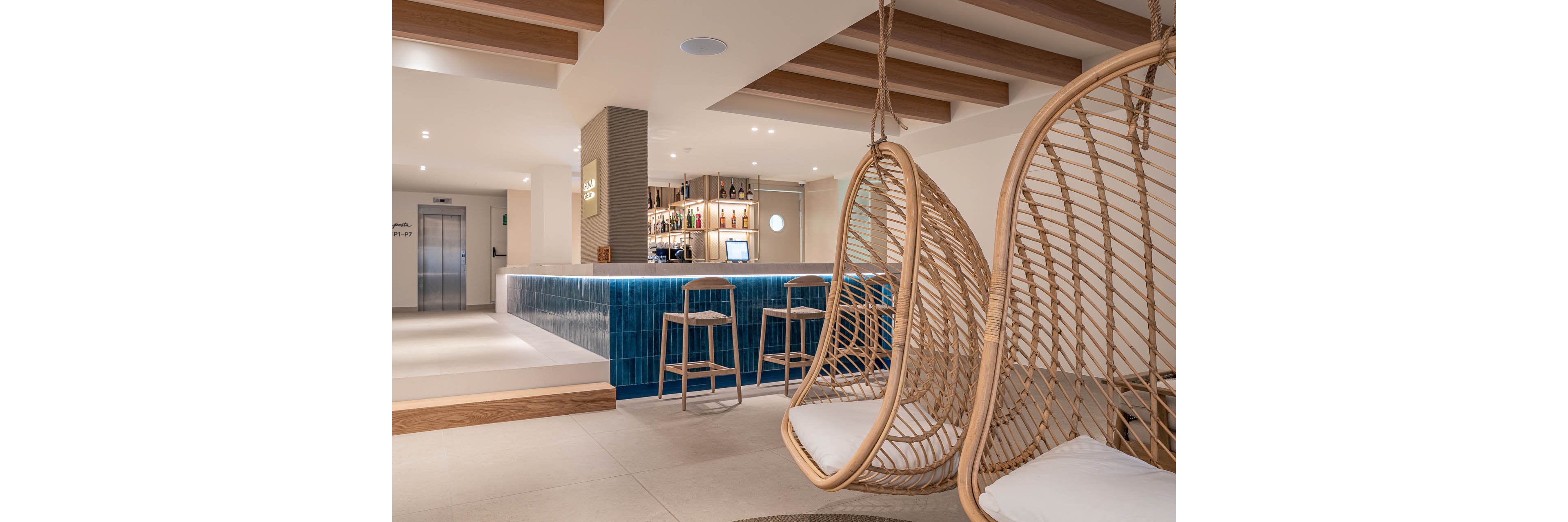 Contemporary bar interior featuring a sleek counter with blue tile facing and wooden bar stools. Two large wicker hanging chairs with white cushions create a relaxed seating area in the foreground. The space is bright and minimal, with neutral walls, light flooring, and exposed wooden ceiling beams. Bottles and glassware are neatly arranged behind the bar, and an elevator is visible in the background, suggesting a hotel or resort setting.