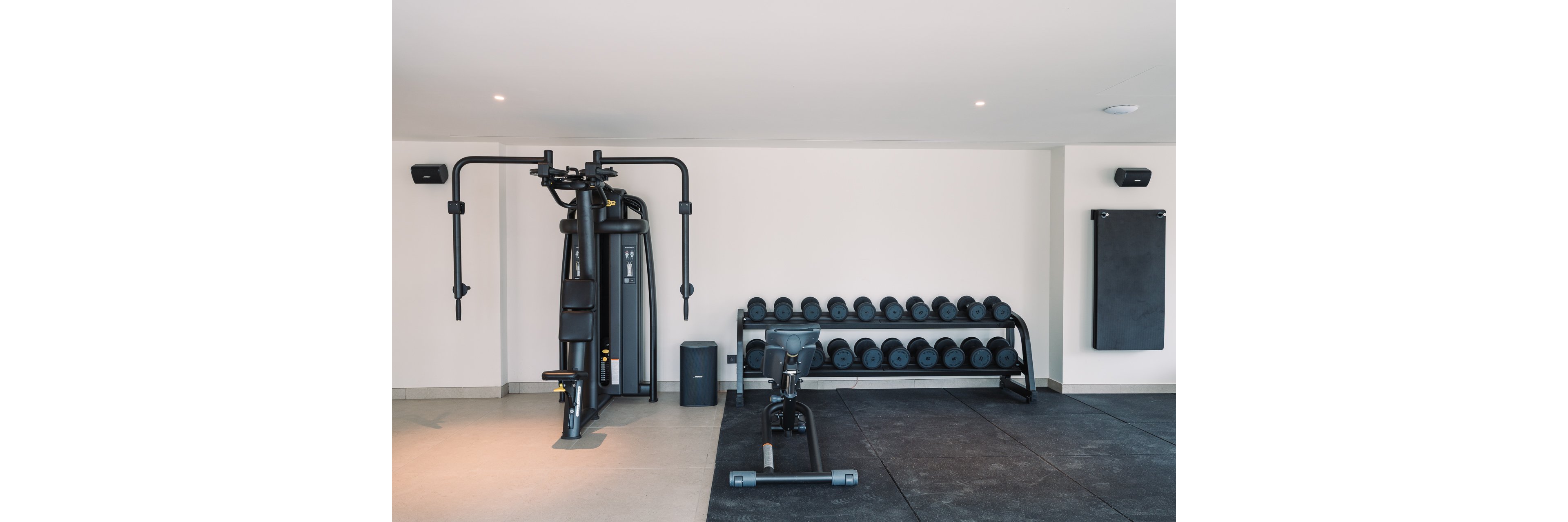 A clean, modern home gym features a multi-station weight machine and a rack of dumbbells against a white wall. A stationary exercise bike is positioned on rubber flooring in the foreground. The space is minimalistic with neutral colors and ample open floor area for workouts. Wall-mounted speakers and a folded exercise mat complete the functional fitness environment.